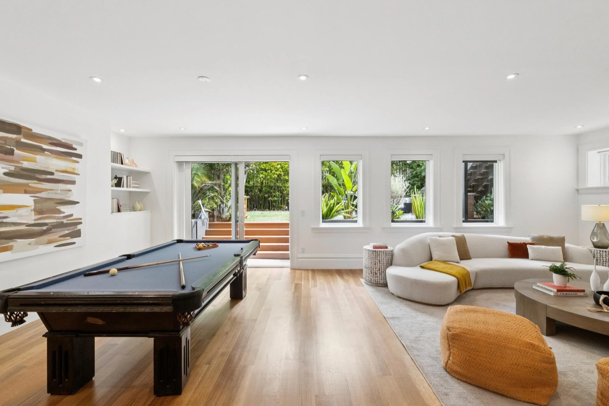 Bright living room with white walls, large windows, a pool table, a curved white sofa with colorful cushions, and a view of a lush green garden outside.