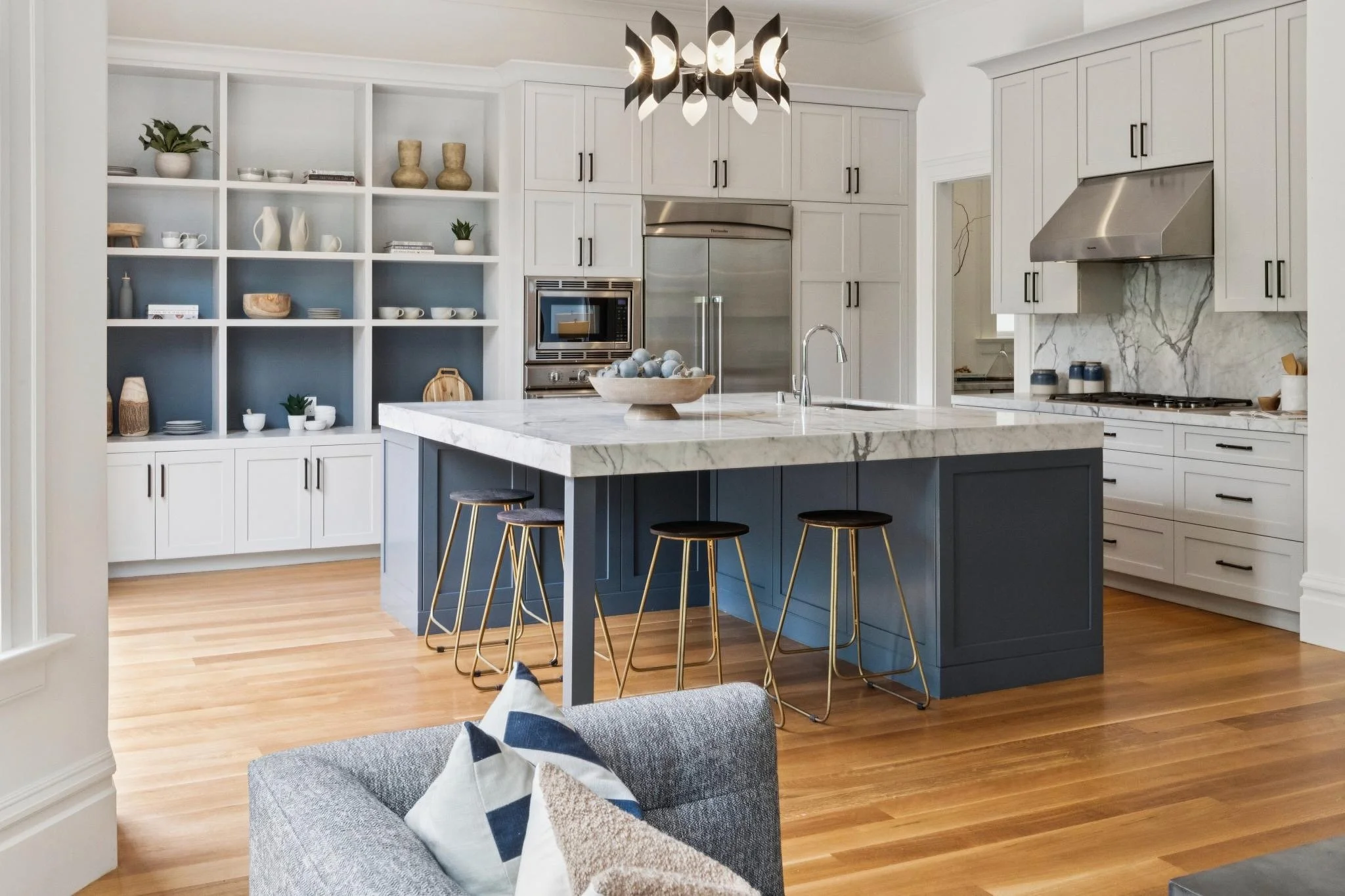 Modern kitchen with white cabinets, marble countertops, a blue island with barstools, open shelving with decor, and a black chandelier
