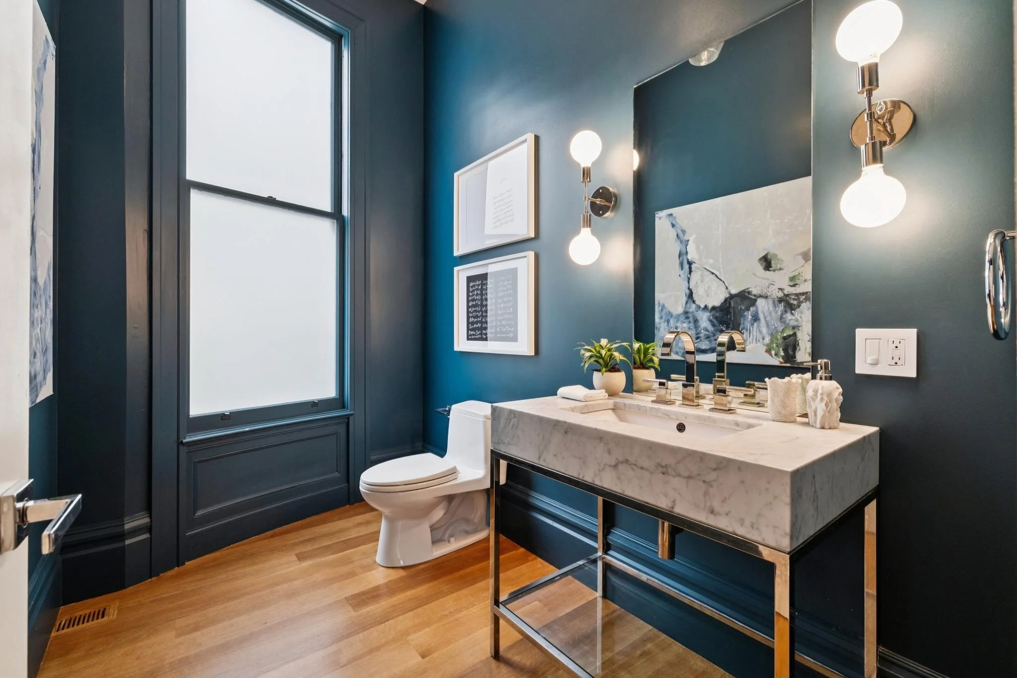 A modern bathroom featuring navy blue walls, a large window, wall-mounted light fixtures, framed artwork, a white toilet, and a white marble sink with two silver faucets. There are decorative plants and art pieces on the sink counter.