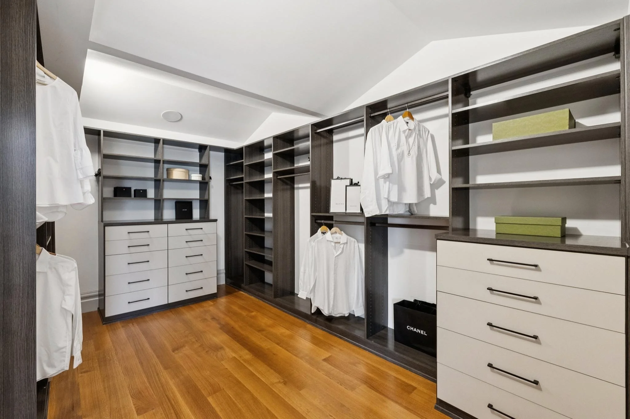 A walk-in closet with dark wood shelving and drawers, white shirts hanging on rods, some boxes on the shelves, and a hardwood floor.