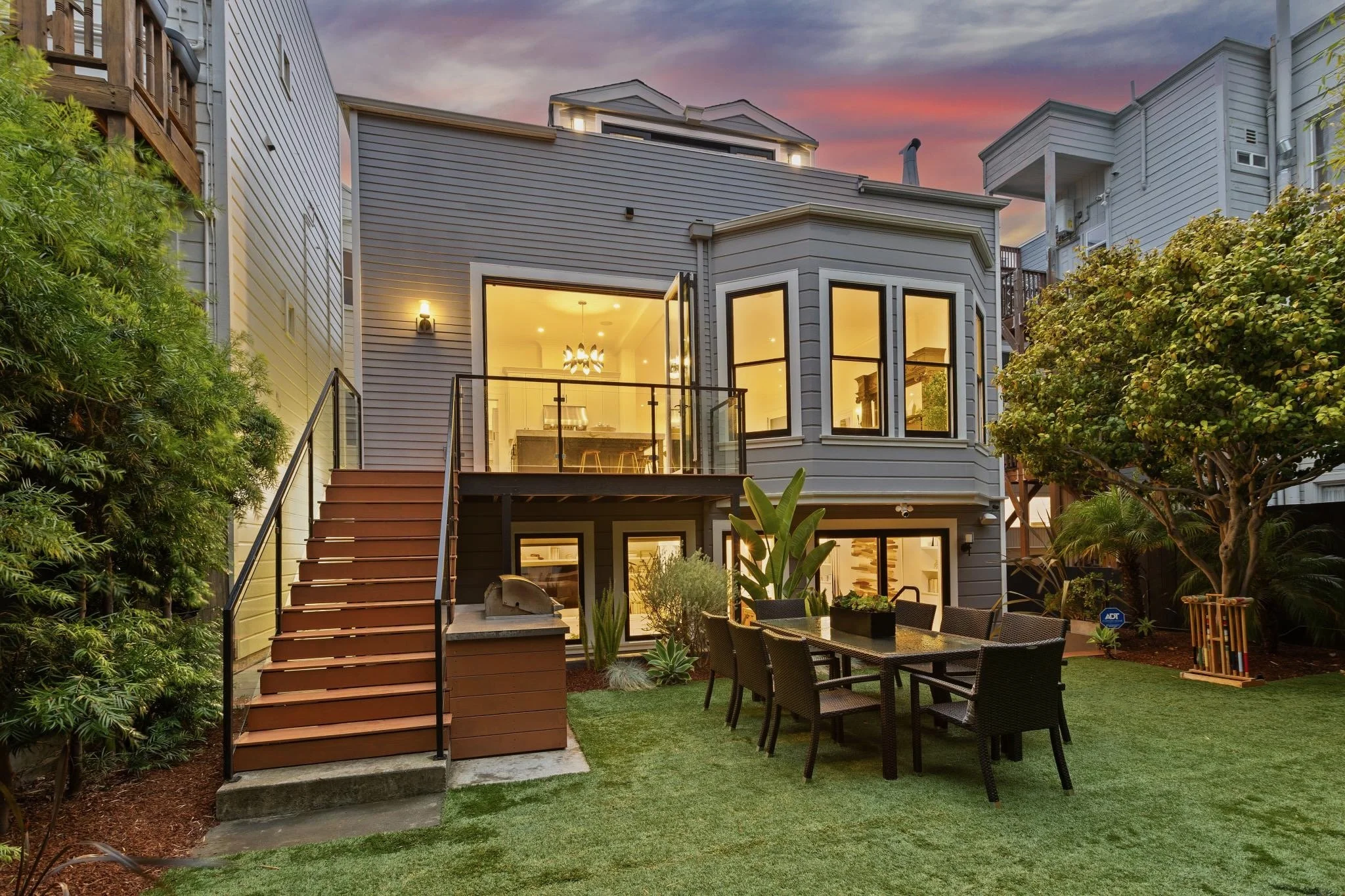 A modern multi-story house with large windows, a small balcony, and a backyard with outdoor dining area, trees, and shrubs at dusk.