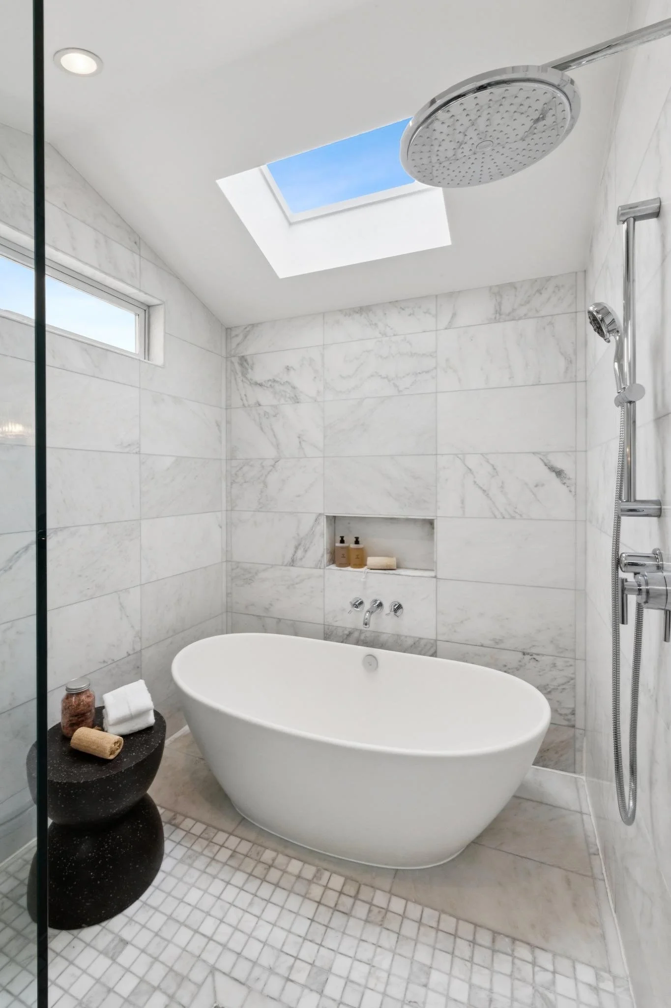 Modern bathroom with white marble walls, a freestanding bathtub, a skylight, a showerhead, and windows for natural light.