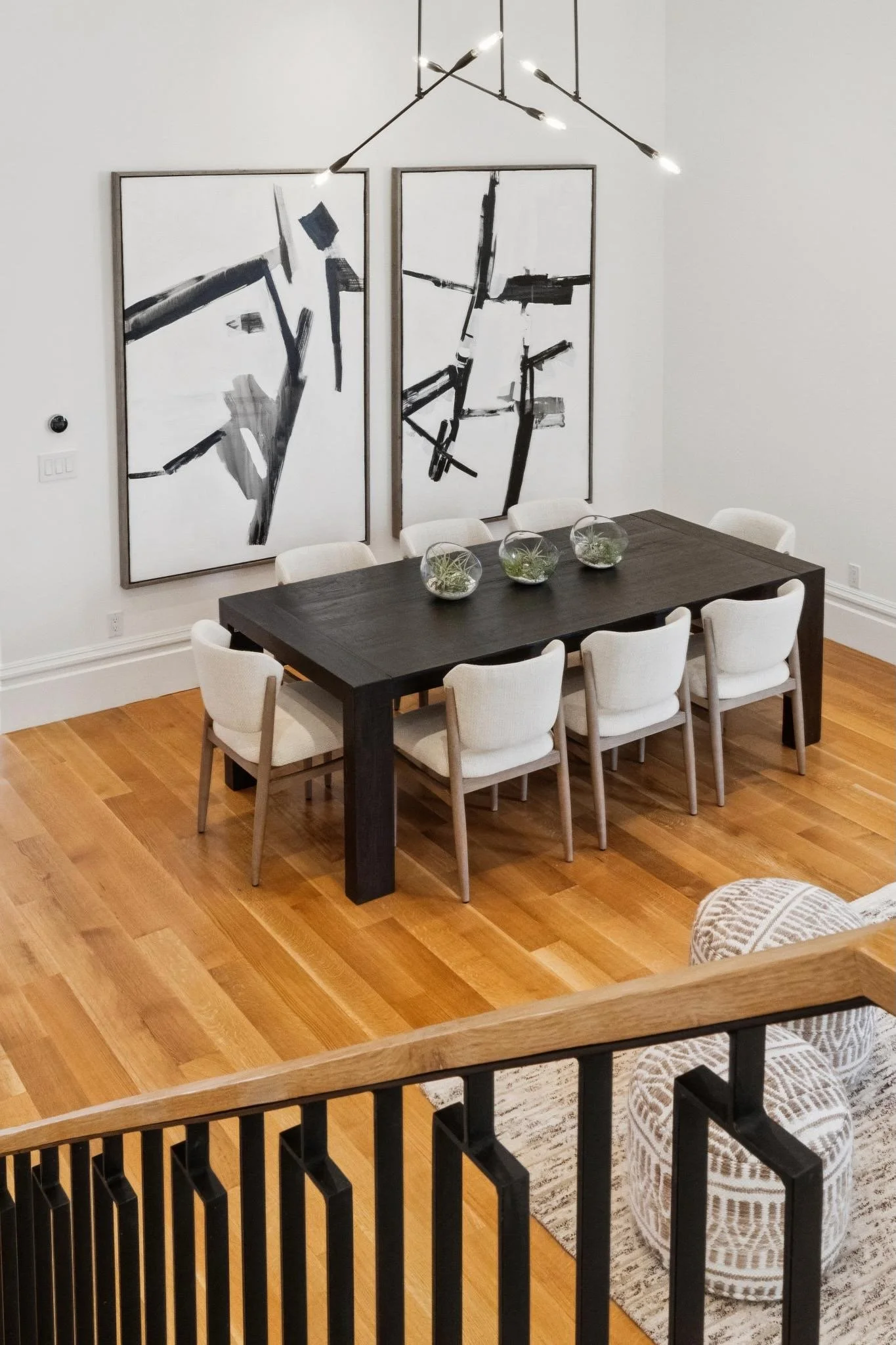 A dining room with a black table surrounded by white chairs, with three glass bowls containing plants on the table. Two large abstract black and white paintings hang on the white wall, and a modern chandelier hangs overhead. The floor is made of warm