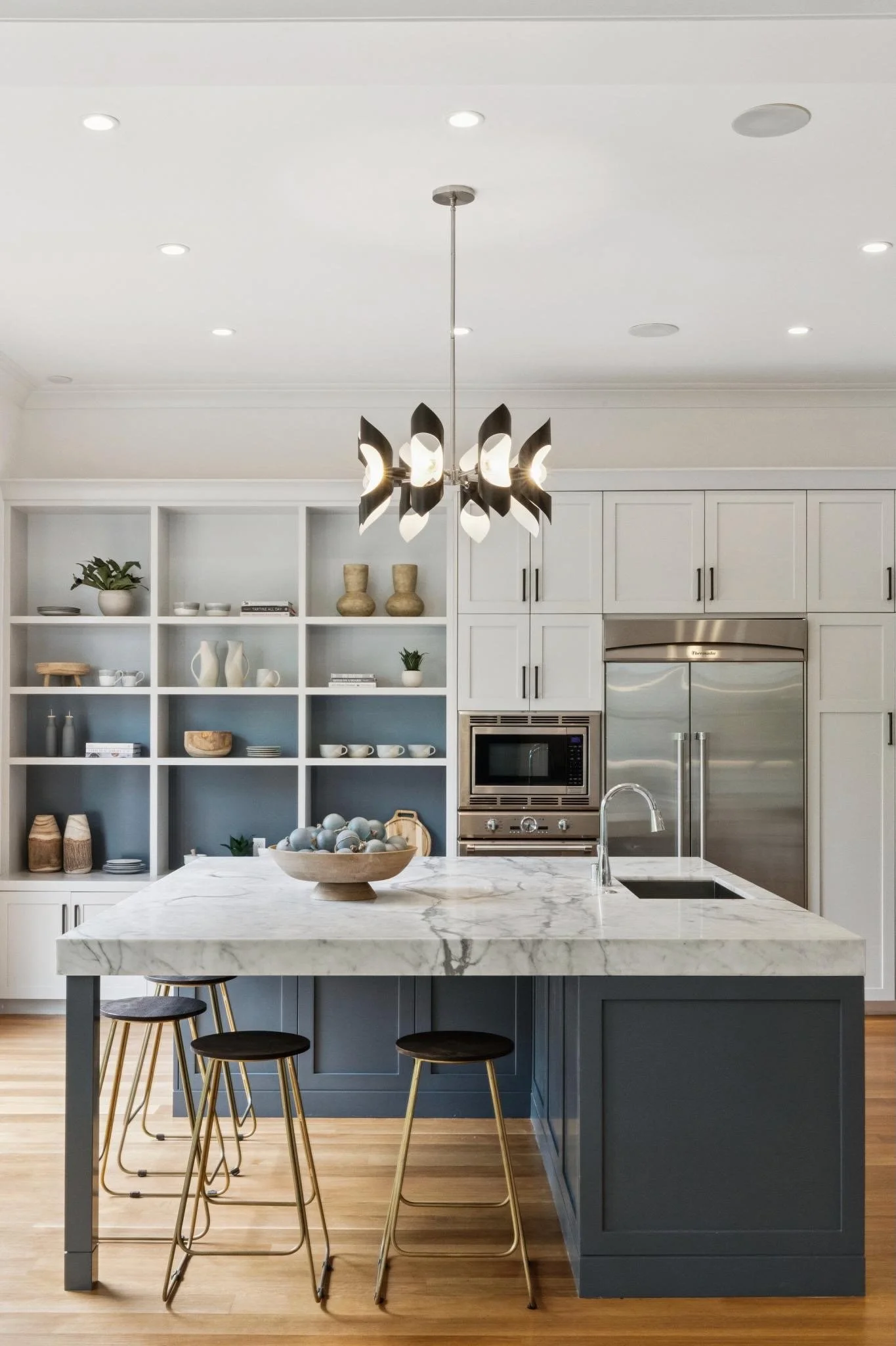 Modern kitchen with white cabinetry, a marble island with seating, stainless steel appliances, and decorative vases on open shelves, illuminated by pendant and recessed lighting.