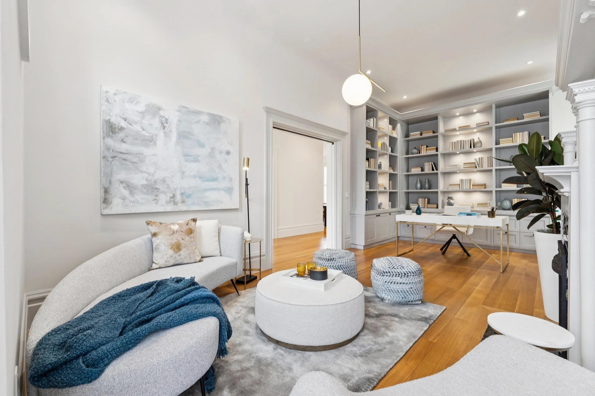 Living room with white sofa, blue throw blanket, and pillows, a round white ottoman, large built-in bookshelves filled with books and decorative items, hardwood floors, and modern lighting fixtures.