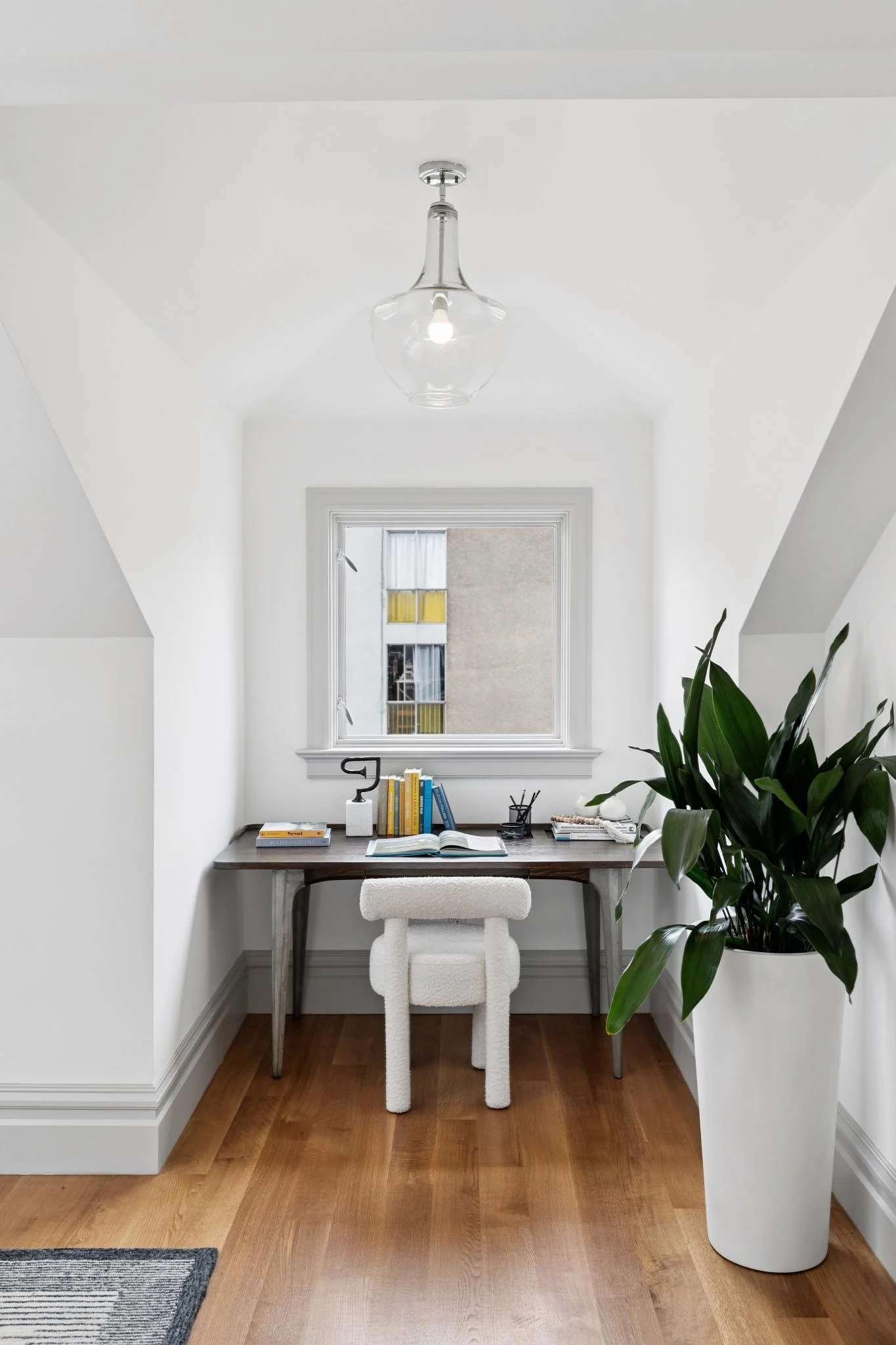 A minimalist home office with a wooden desk, books, and a white fluffy chair. A large potted green plant is on the right. A window with a view of an apartment building is above the desk. A modern glass pendant light hangs from the ceiling.