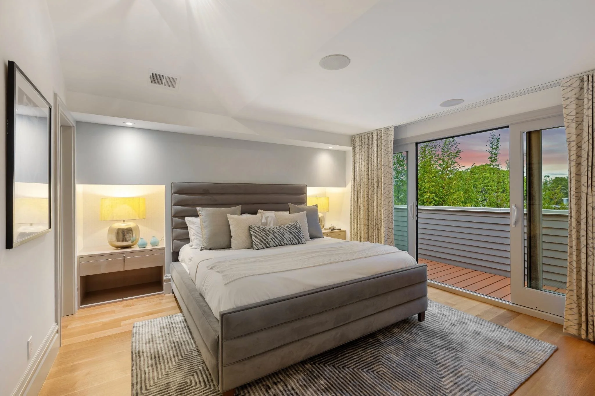 A modern bedroom with a large bed, gray upholstered headboard, multiple pillows, bedside table with a yellow lamp, and sliding glass doors leading to a balcony with greenery and a sunset sky visible outside.