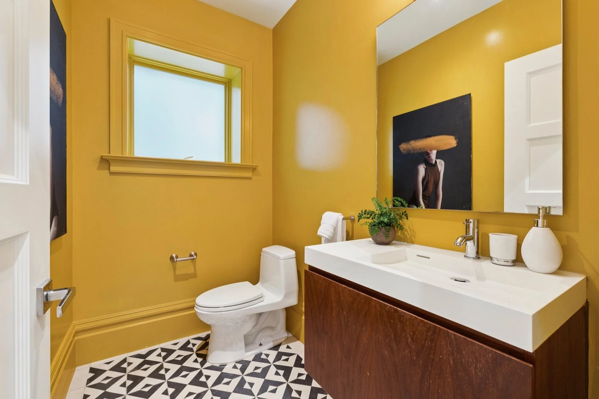 A bathroom with yellow walls, a window, a white toilet, a wooden vanity with a white countertop, a large mirror, a potted plant, and decorative black and white patterned floor tiles.