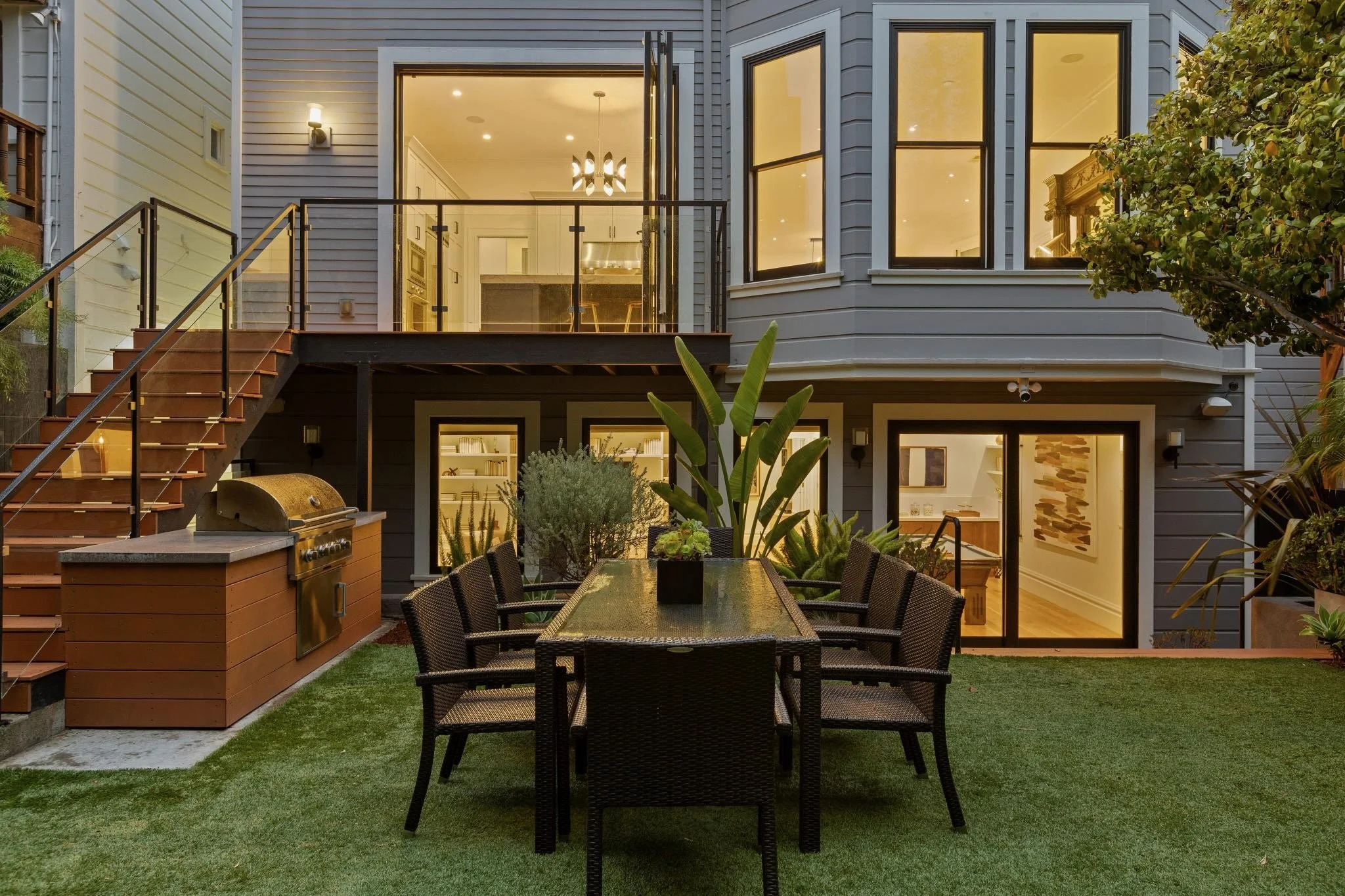 Outdoor patio area with a dining table and chairs, built-in grill, and view into a modern house with large windows and interior lighting.