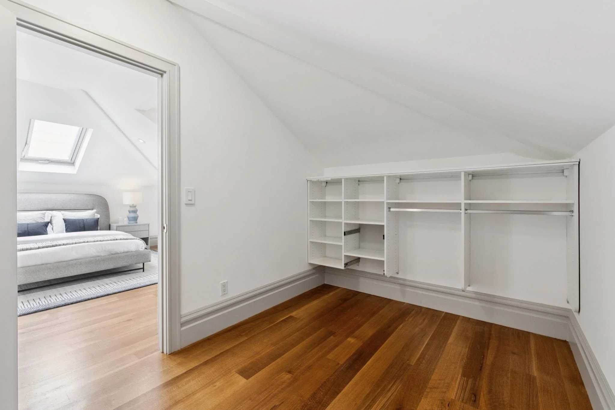 Unfinished walk-in closet with white shelving and a sloped ceiling, hardwood floors, and an open doorway leading to a bedroom with a bed, nightstand, lamp, and skylight.