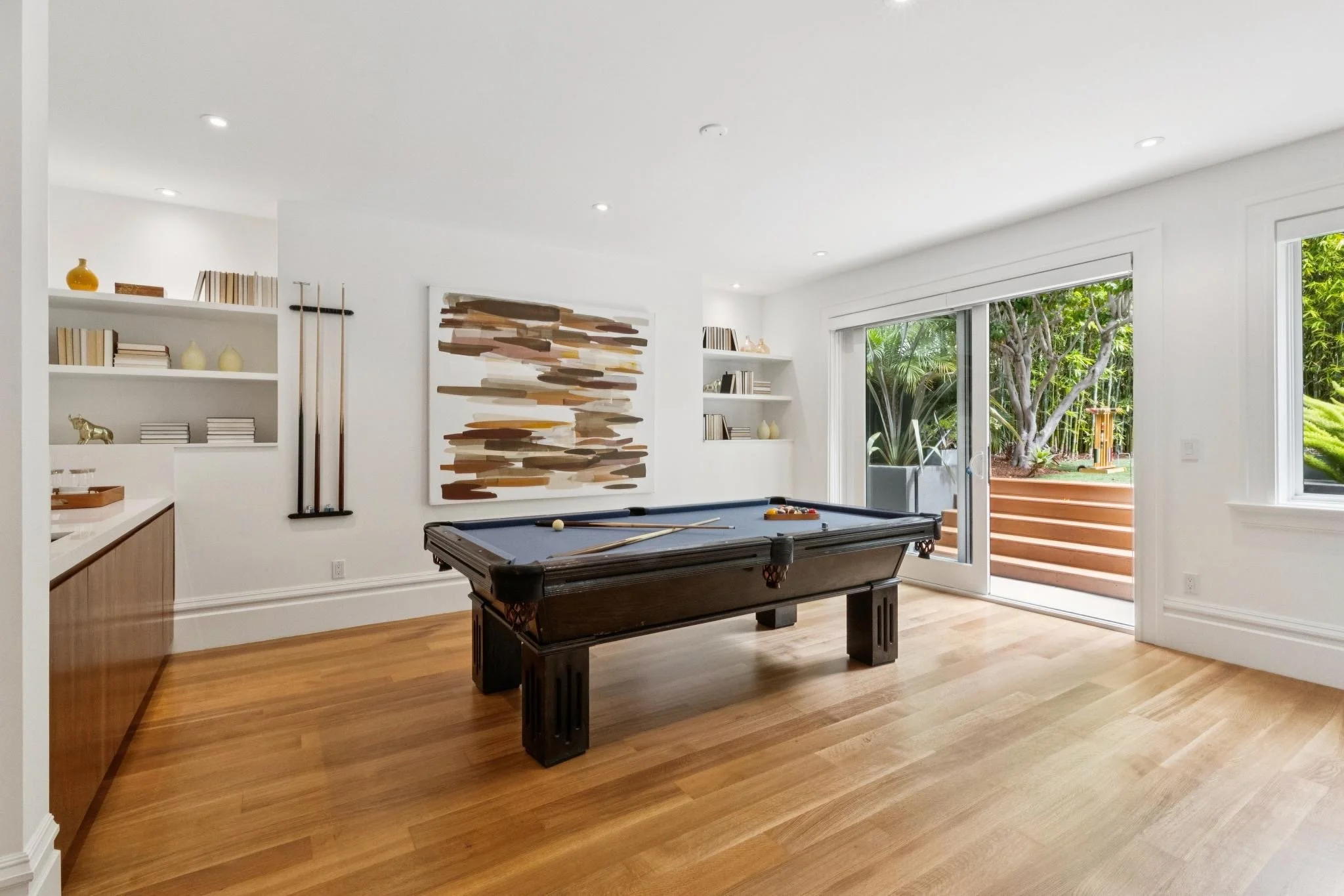 Living room with sliding glass door opening to a backyard with trees. Contains a pool table, built-in shelves with books and decorative items, and artwork on the wall. Hardwood floors and white walls.