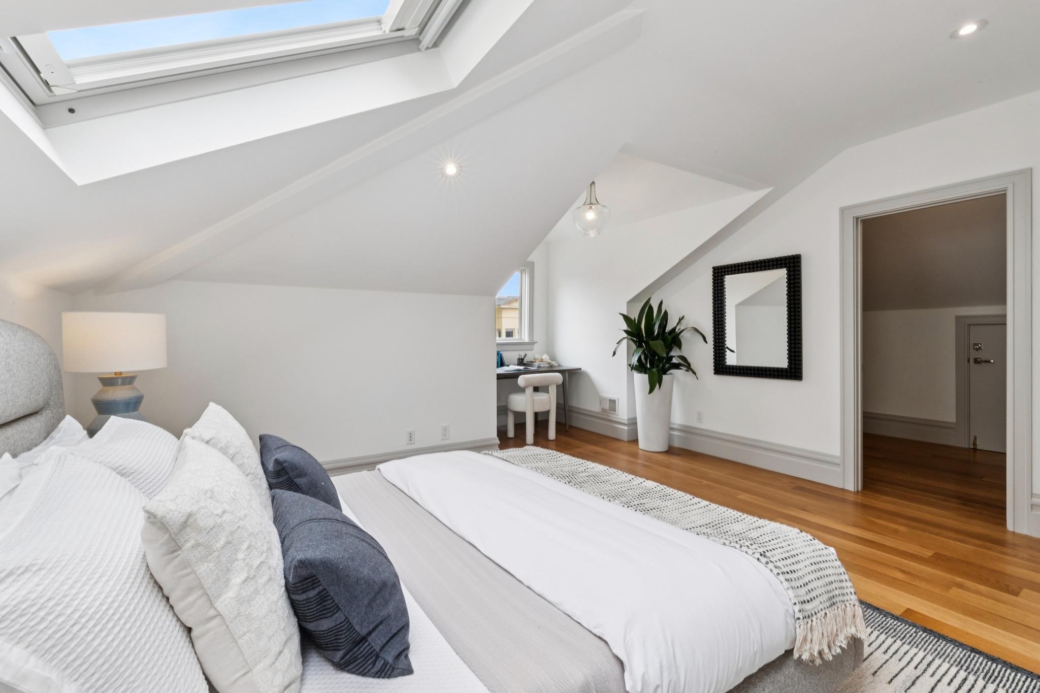 A bedroom with white walls, a sloped ceiling, and a large skylight window. There is a bed with white bedding and pillows, a nightstand with a lamp, a small desk with a chair under a window, a large potted plant, a black framed mirror, and wood floori