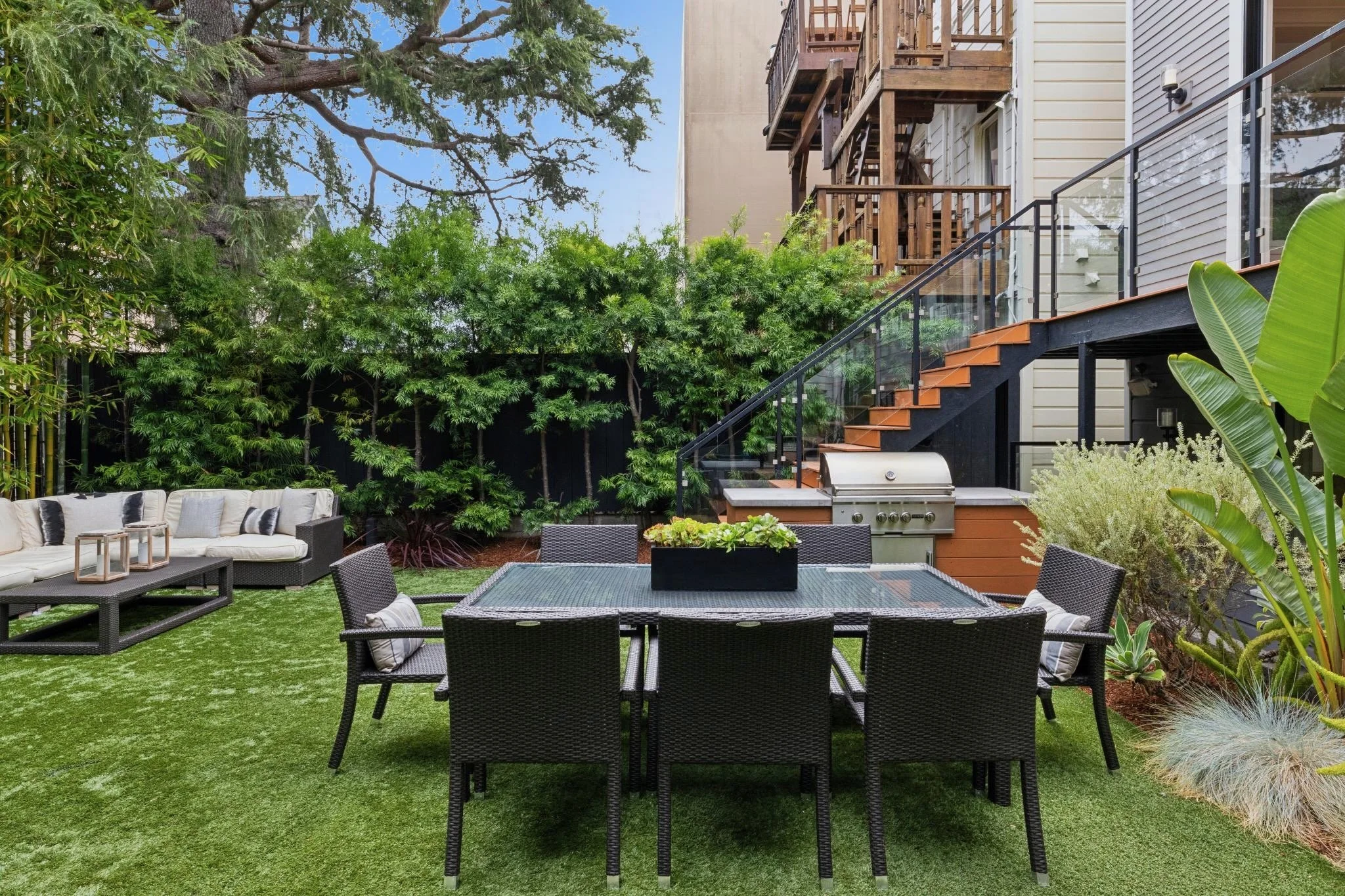 A backyard patio with outdoor seating, a dining table, a built-in grill, surrounded by green trees and plants, with a multi-story house featuring a wooden staircase and deck.