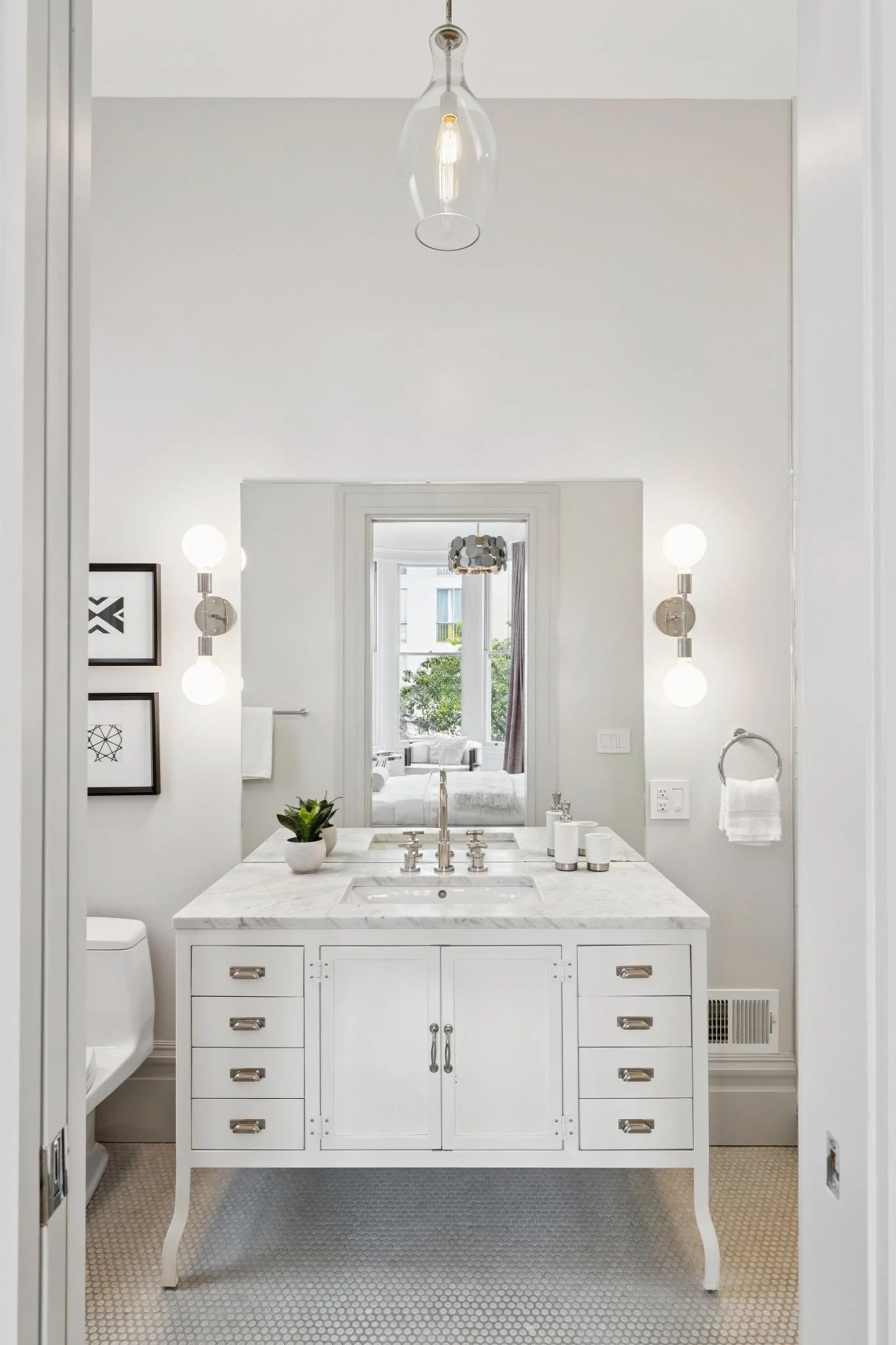 A white bathroom vanity with a marble countertop, silver handles, and a mirror above it, illuminated by two wall-mounted lights. There is a small potted plant on the counter, and a towel hanging on a rack. The bathroom has white walls and windows in 