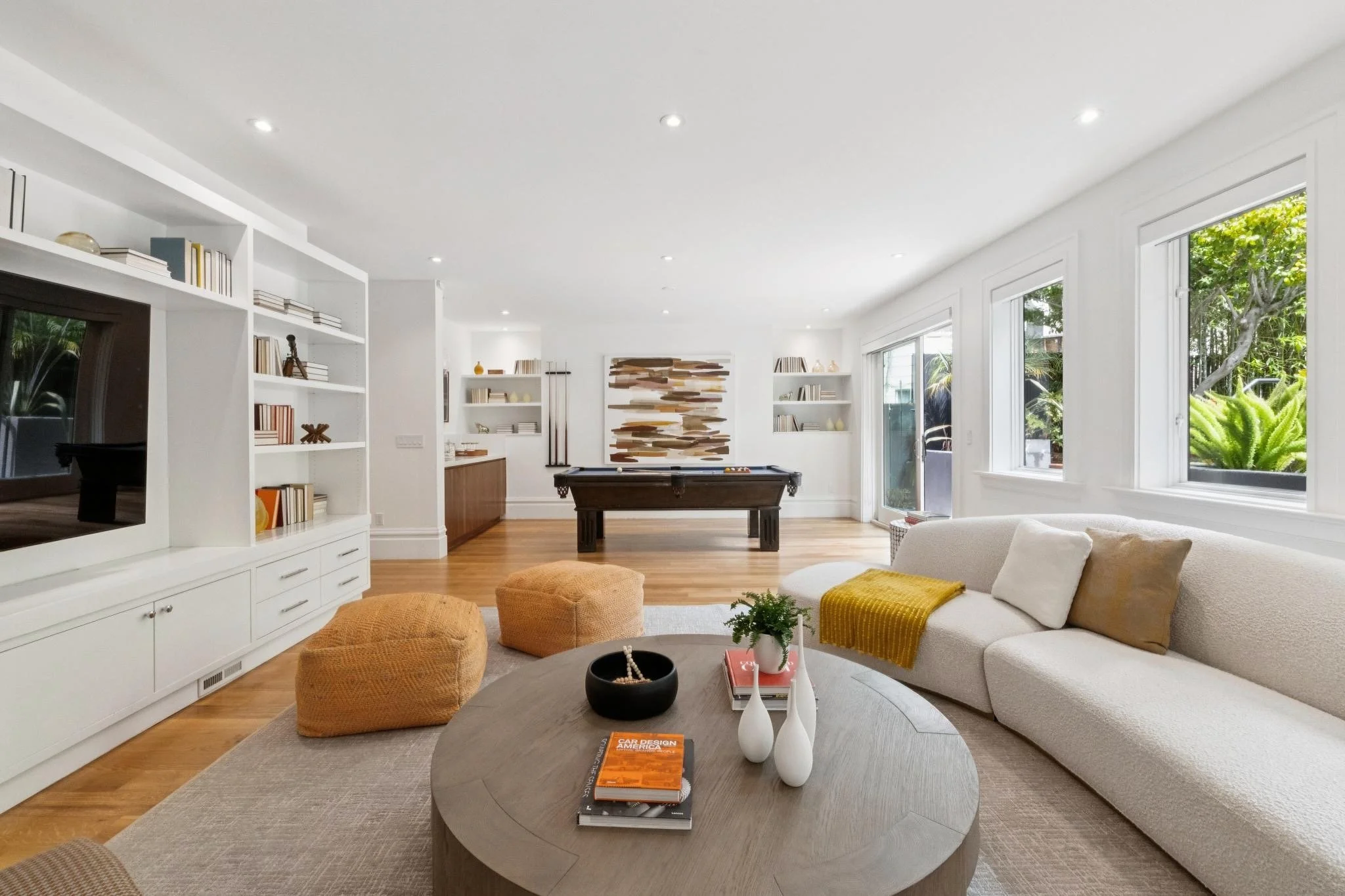 Bright living room with white walls and ceiling, large windows showing green outdoor trees, beige sofa with pillows, a round coffee table with books and decorative items, built-in white shelves with books and decor, pool table, and hardwood floors.