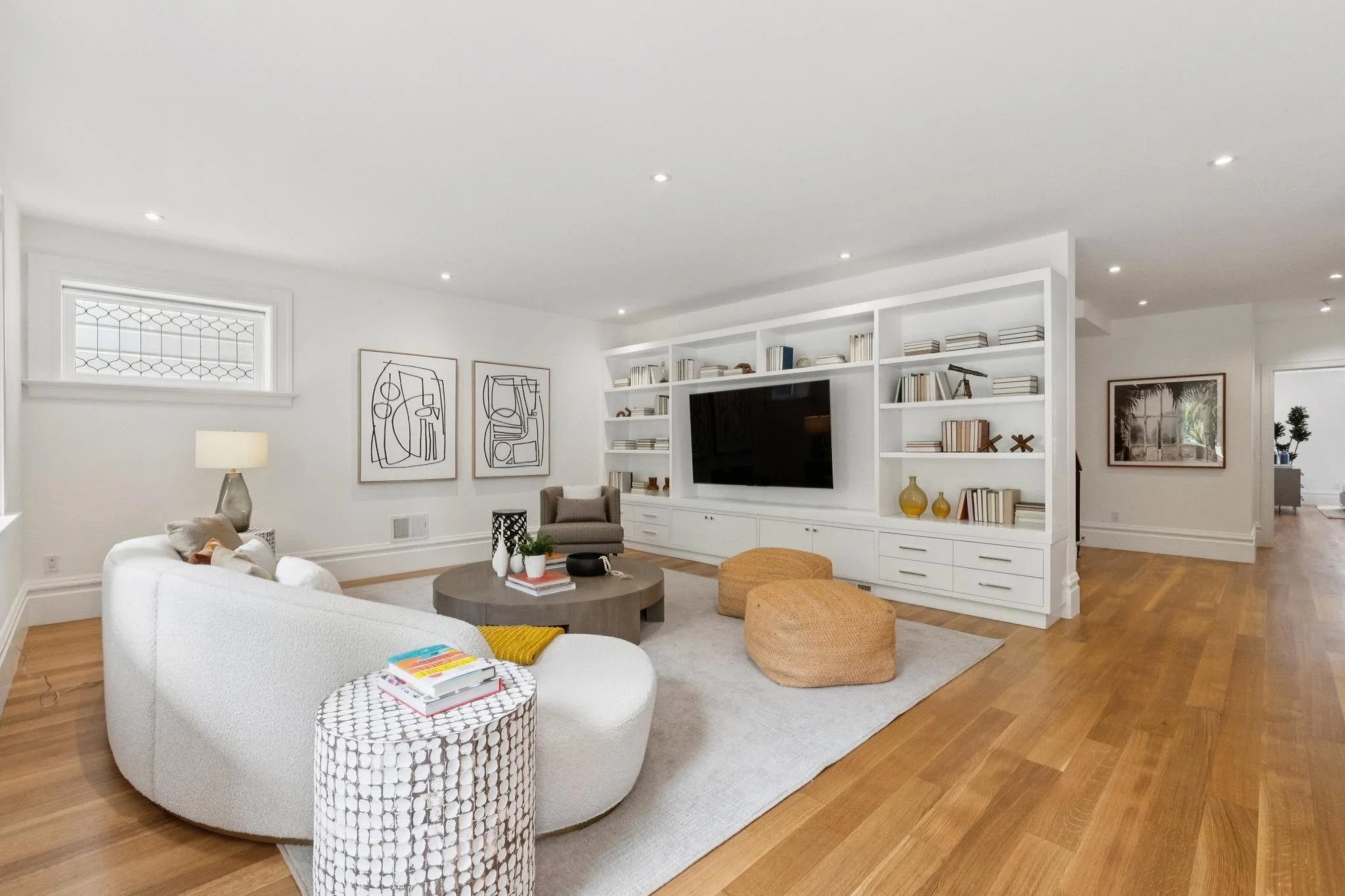 Modern living room with white walls, hardwood floors, a white sofa, two orange poufs, a round coffee table with books and decorative items, built-in white bookshelf with books and decor, framed artwork on the wall, and large windows with natural ligh
