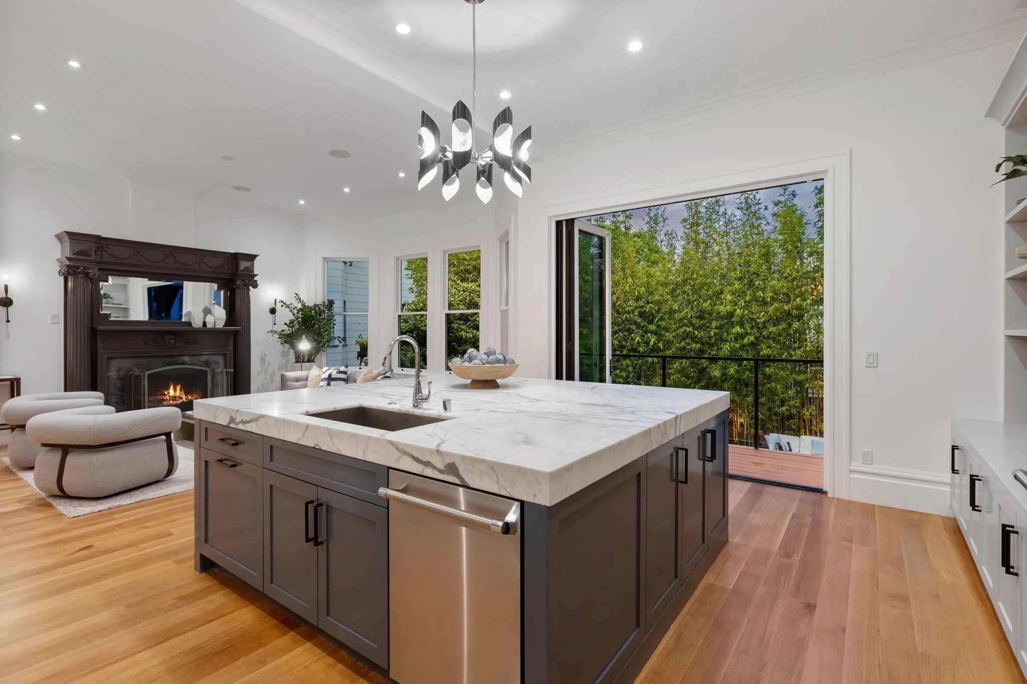 Modern open-concept kitchen with a large marble island, black cabinets, hardwood floors, and large windows overlooking greenery. Rest of the living space includes a seating area with cream-colored chairs and a fireplace with an ornate dark wood mante