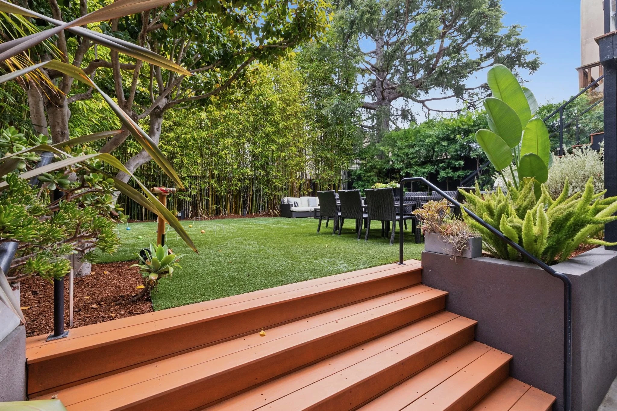 Backyard patio with wooden stairs leading to a lawn surrounded by various green plants and trees, including banana plants and tall pines, with outdoor dining furniture and a cushioned outdoor sofa.