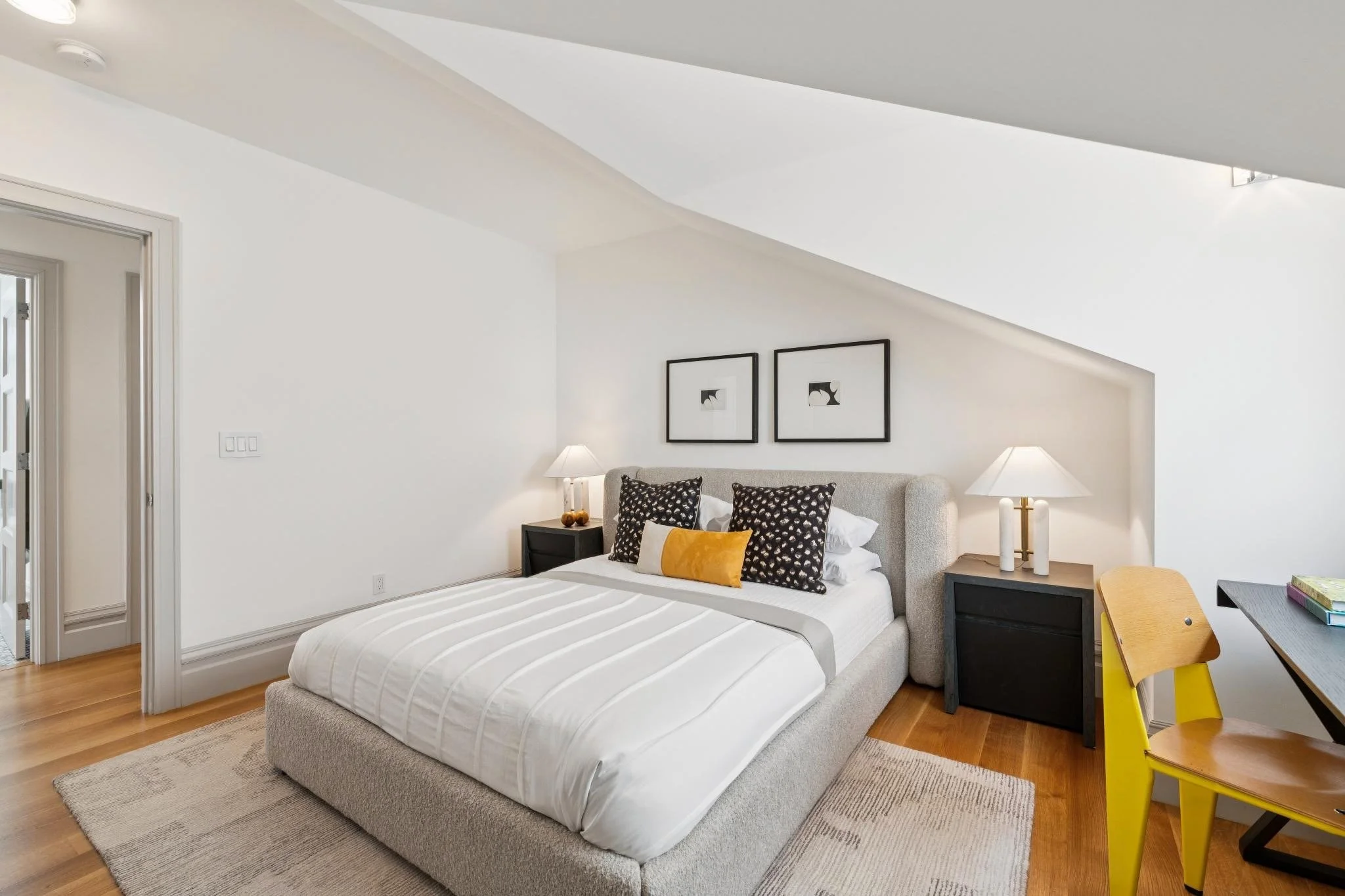 Modern bedroom with white walls, a bed with a beige upholstered headboard, black and white patterned pillows, a mustard yellow accent pillow, and a white striped bedspread. Flanking the bed are black nightstands with white lamps. Two black-framed abs