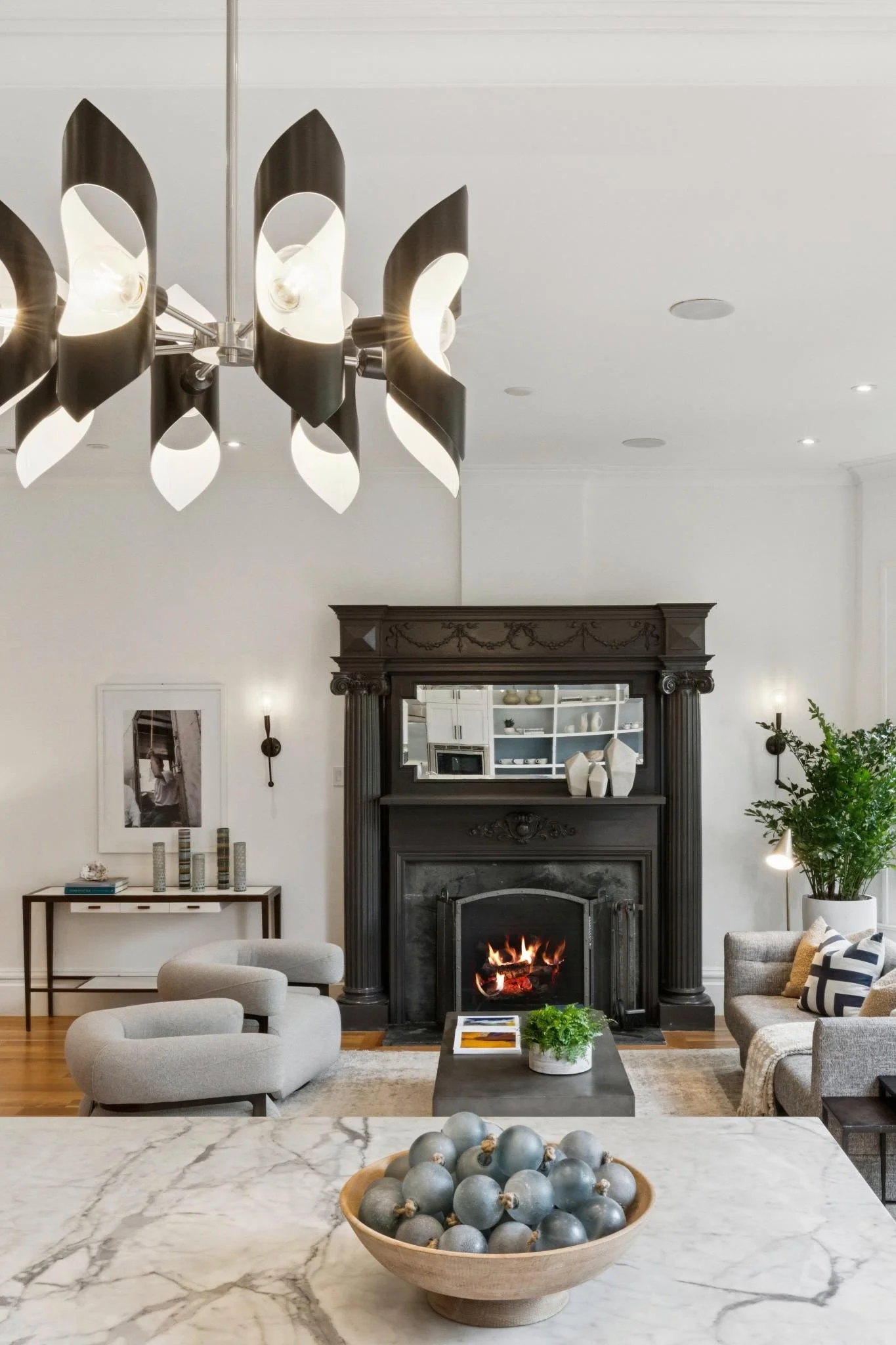 Living room with fireplace, modern chandelier, and neutral furniture including a gray armchair and a gray sofa, with a marble coffee table and decorative items.