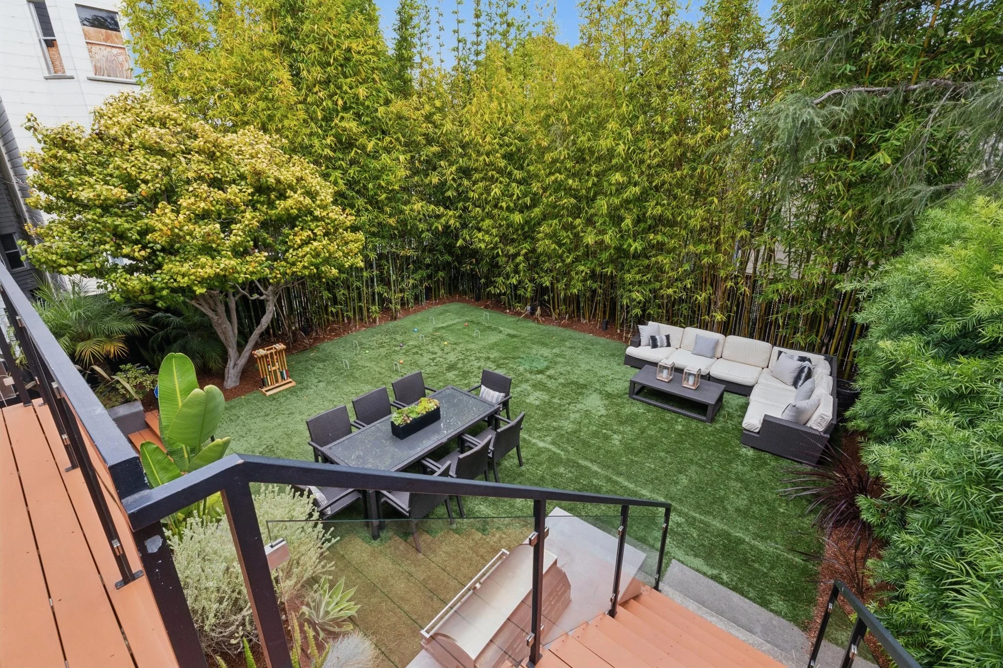 A backyard patio with black outdoor dining set, a beige outdoor sectional sofa, and lush greenery including tall bamboo, trees, and potted plants. There is artificial grass on the ground and a small outdoor game with balls and hoops.