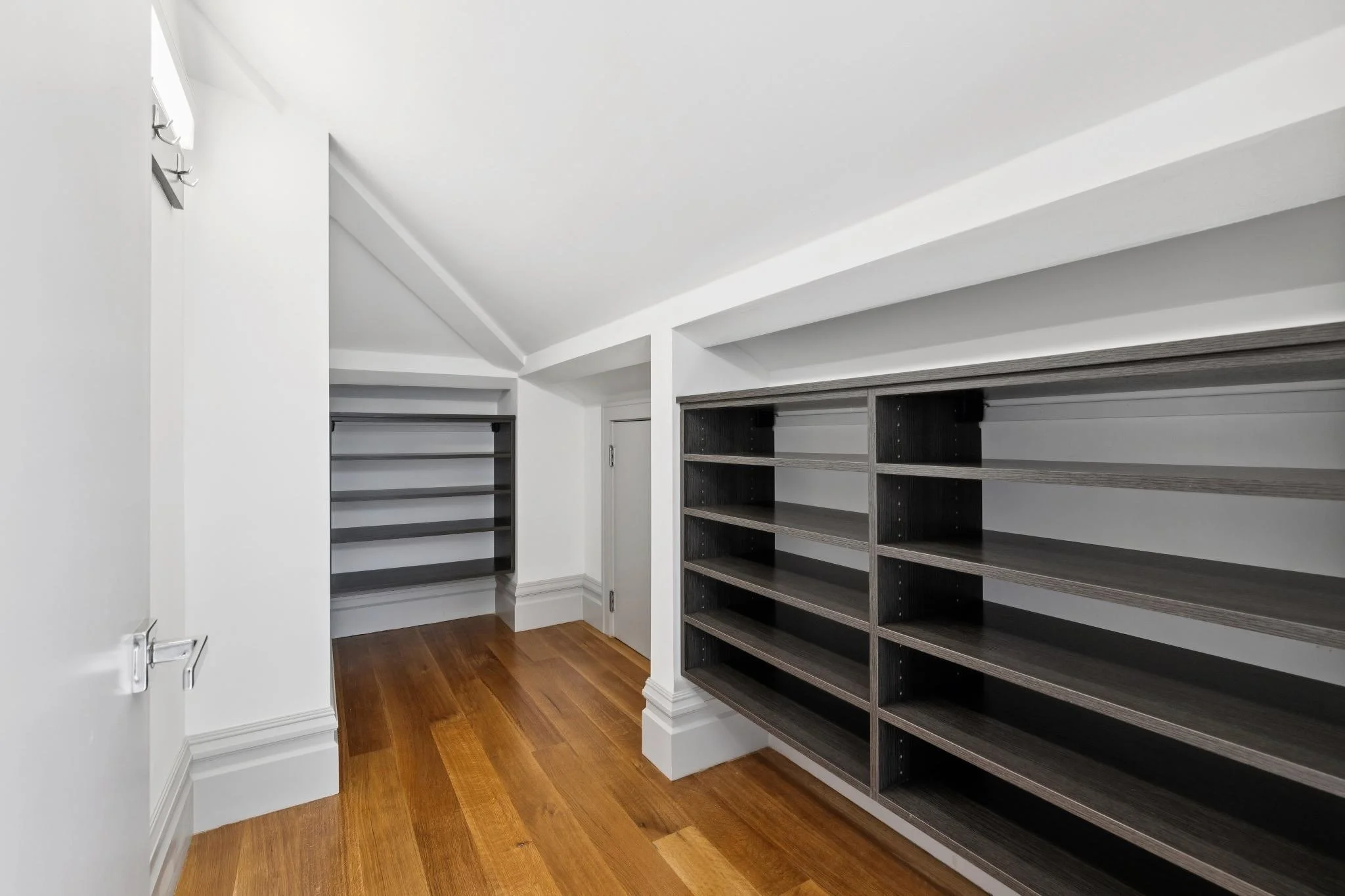 Empty walk-in closet with white walls and dark wood shelves, hardwood floors, and a small window.