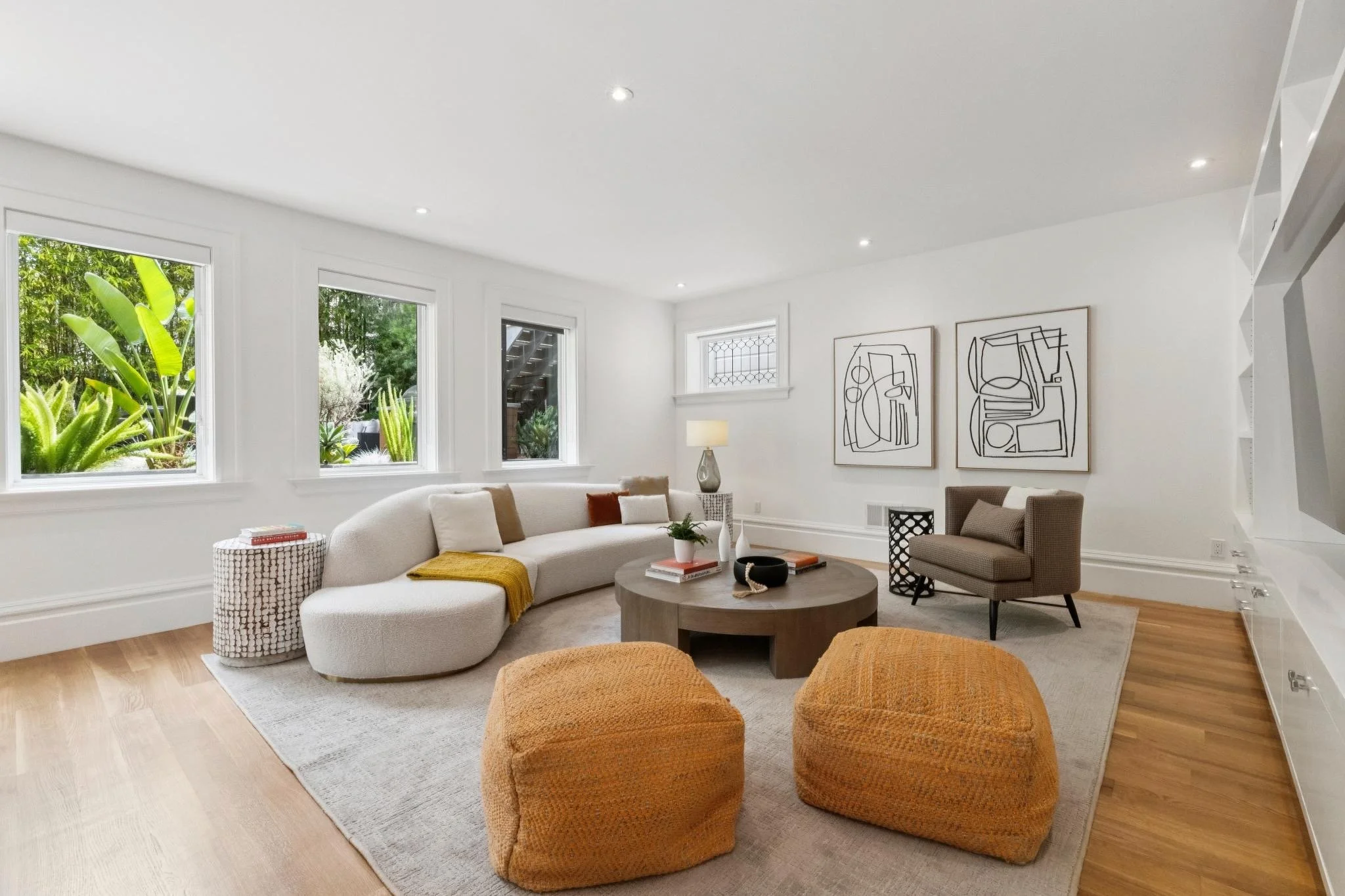 Bright living room with white walls, large windows showing greenery outside, modern abstract art on the walls, and a variety of seating including a curved sofa and orange ottomans.
