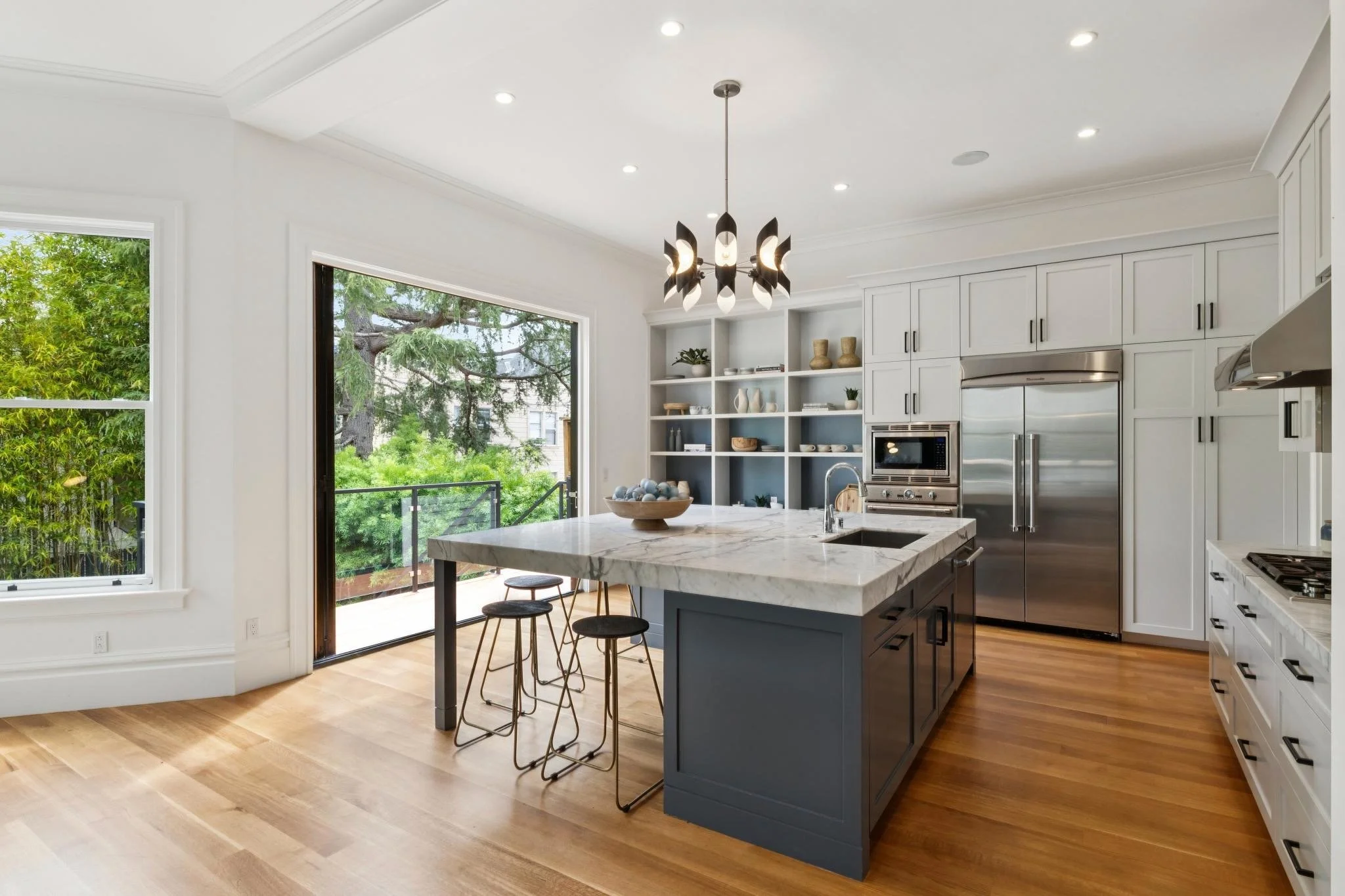 Modern kitchen with white cabinets, stainless steel appliances, marble island, wooden floor, large windows, and leafy trees outside.