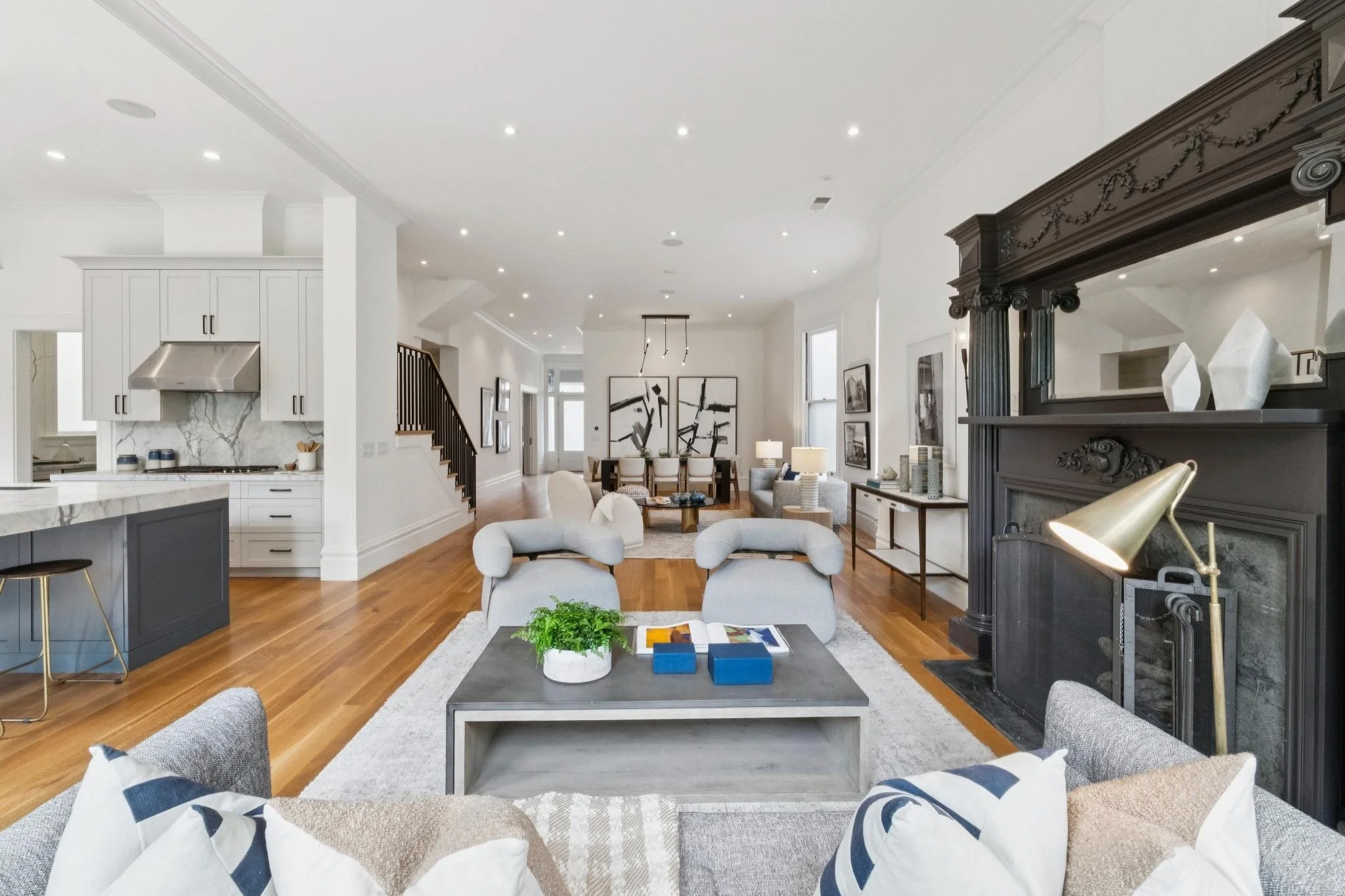 Open-concept living room and kitchen with white walls and ceiling, hardwood floors, modern furniture, and a dark fireplace mantle.
