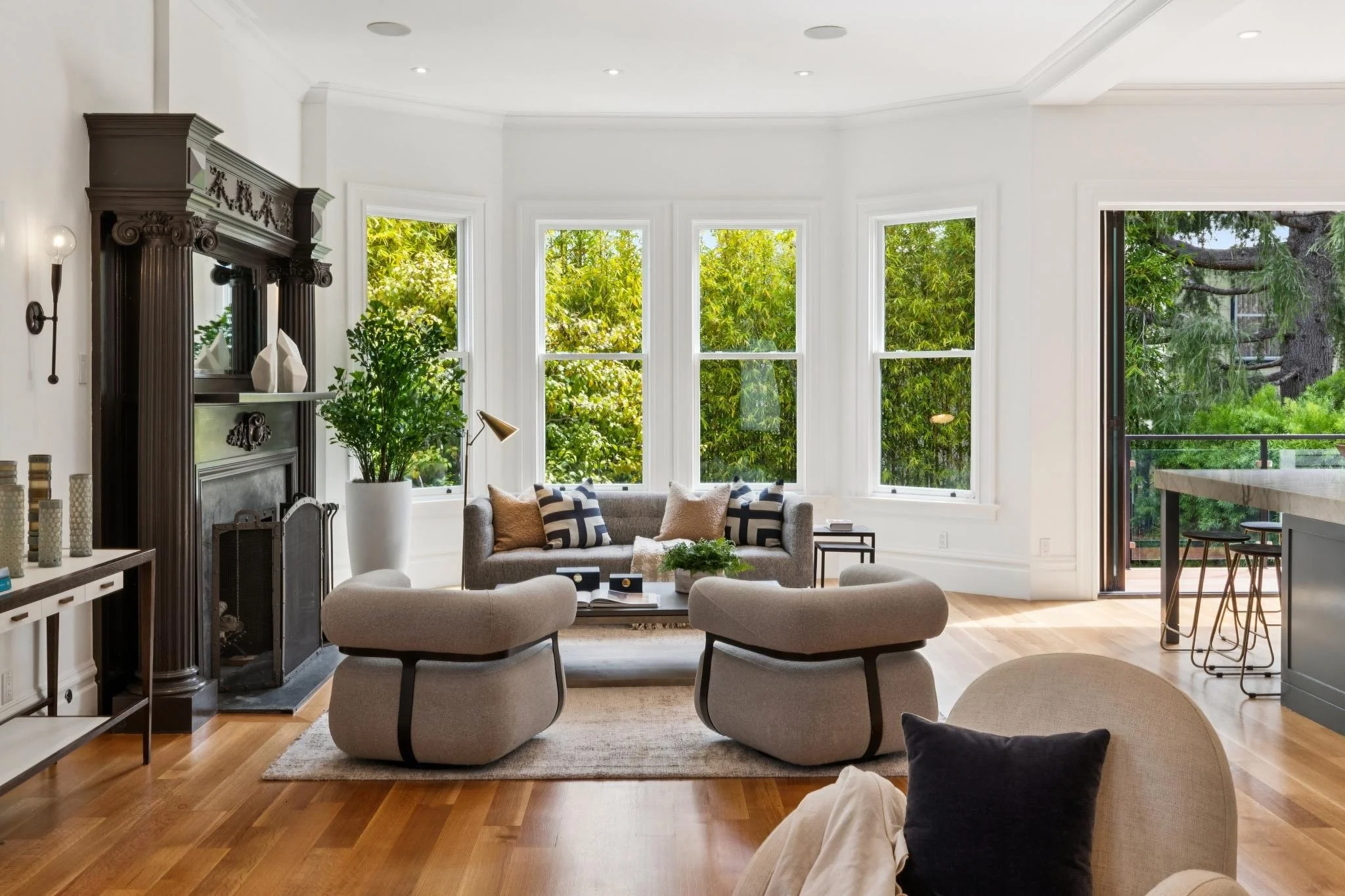 Modern living room with large bay windows showing green trees outside, featuring a gray sofa with pillows, a coffee table, and a fireplace with decorative items.