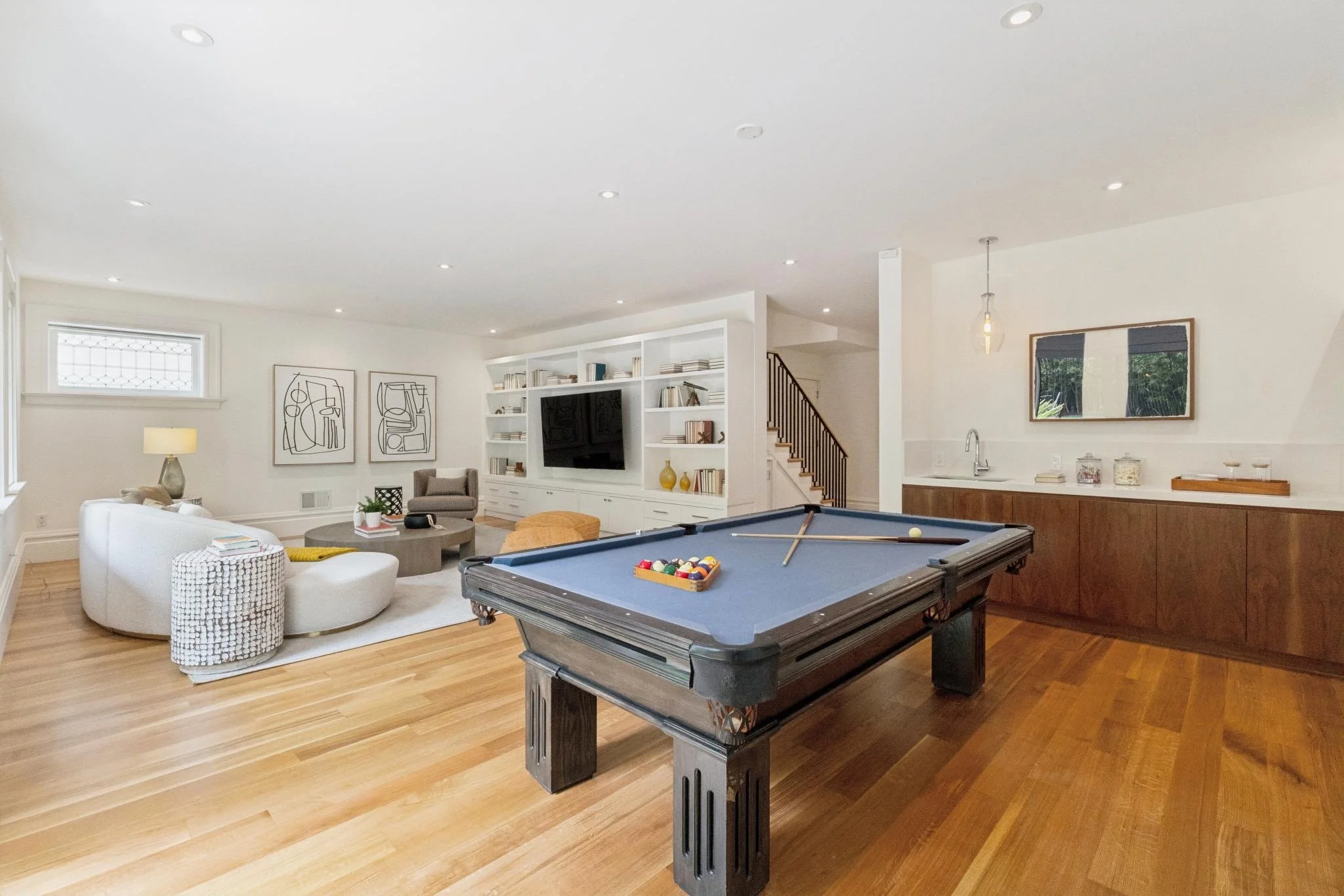 Living room with white walls, hardwood flooring, a pool table, white bookshelf, wall art, and a kitchen area.