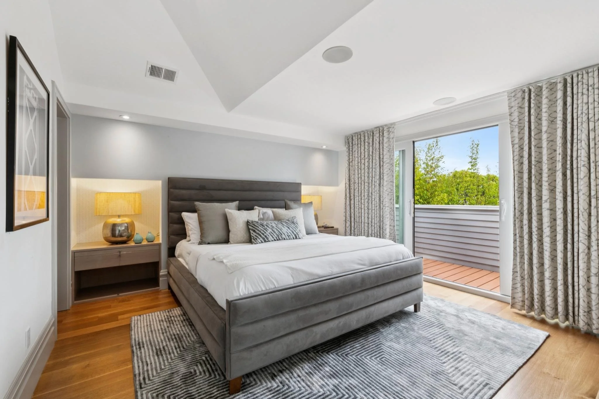 Modern bedroom with a large bed, gray upholstered headboard, decorative pillows, bedside table with a lamp, and a large sliding door with patterned curtains leading to a balcony with greenery outside.