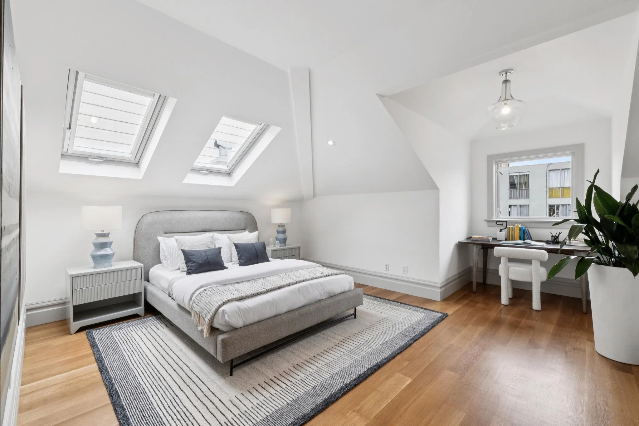 Modern bedroom with sloped white ceiling, two skylight windows, a gray upholstered bed with white bedding and black pillows, white nightstands with lamps, a wooden desk with a plant and books, hardwood floor, and a window with a city view.