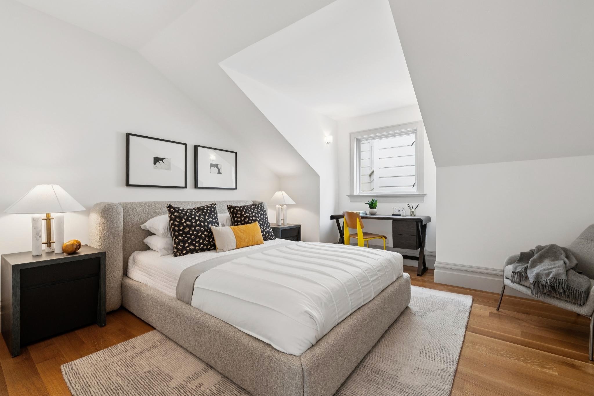 A bedroom with a beige upholstered bed, black nightstands, two white lamps, black and white artwork, a small desk with a yellow chair, a window, and hardwood floors.
