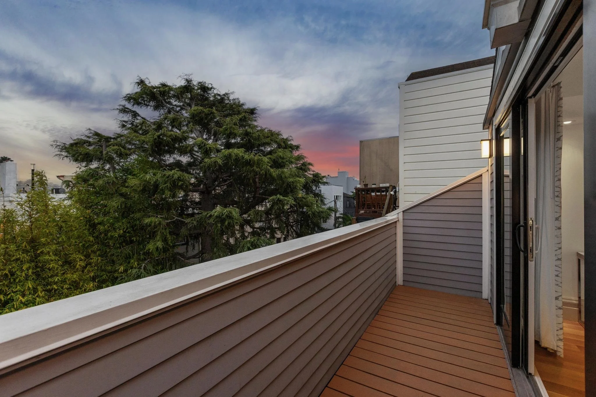 View from a balcony showing the sky at dusk, a large tree, and neighboring buildings with fencing and outdoor furniture.