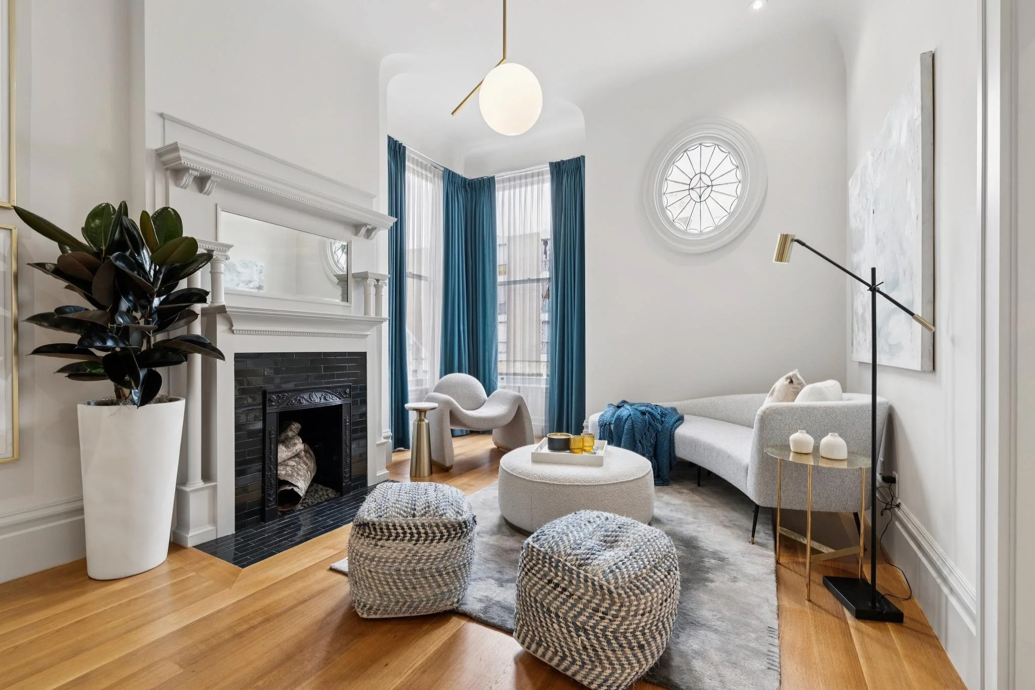 Living room with white walls, a black fireplace, blue curtains, a white sectional sofa, a gray armchair, multiple textured poufs, and modern decor elements such as a large potted plant, a gold side table, and a floor lamp.