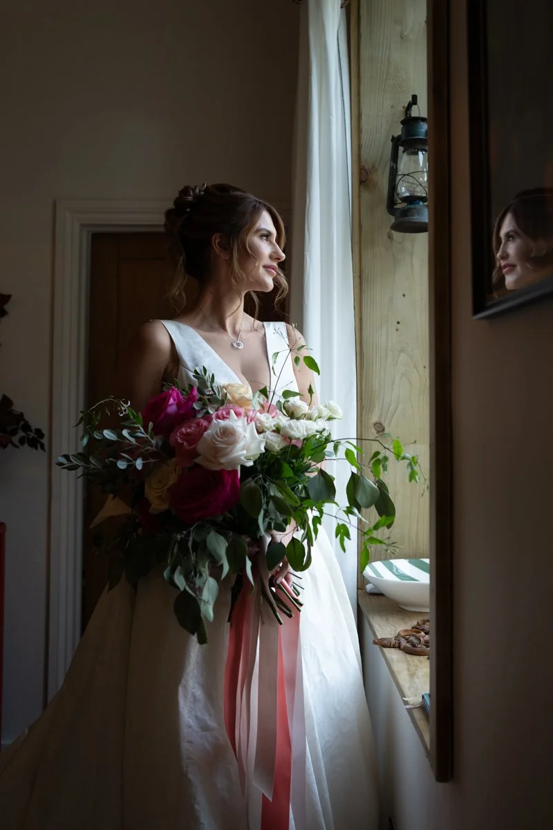 A bride with brown hair styled in an updo wearing a white wedding dress, holding a bouquet of pink and white roses with green foliage, standing by a window with white curtains inside a rustic room with wood accents, looking at her reflection in a mirror.