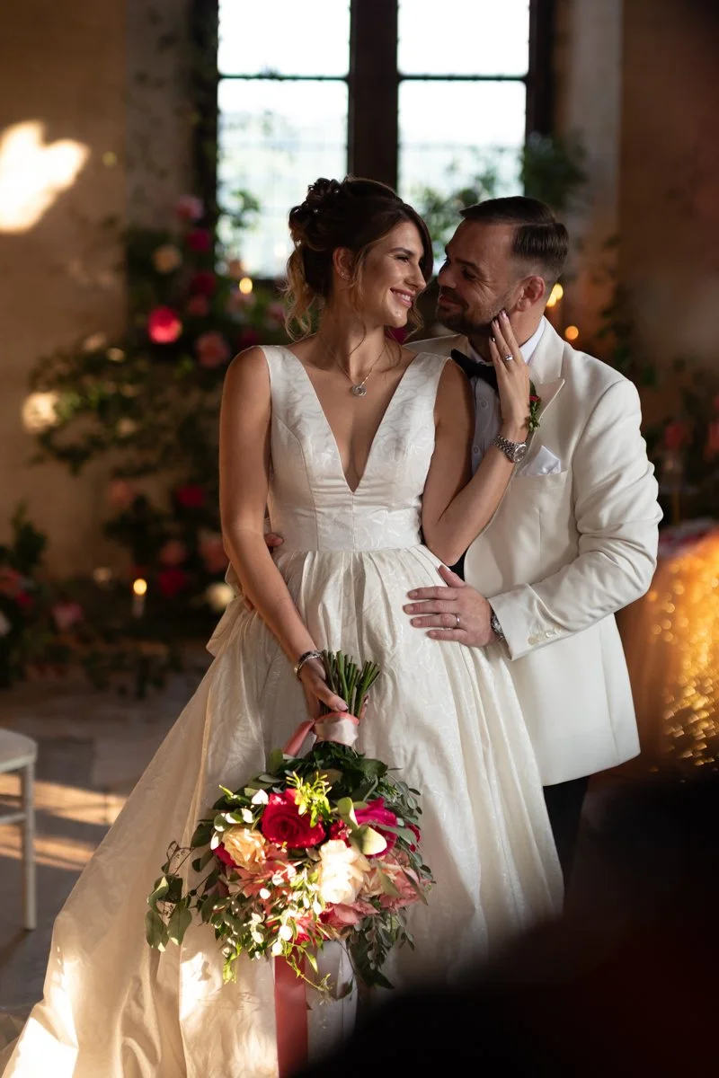 A bride and groom sharing a moment indoors, with the bride holding a bouquet of flowers and both smiling at each other.