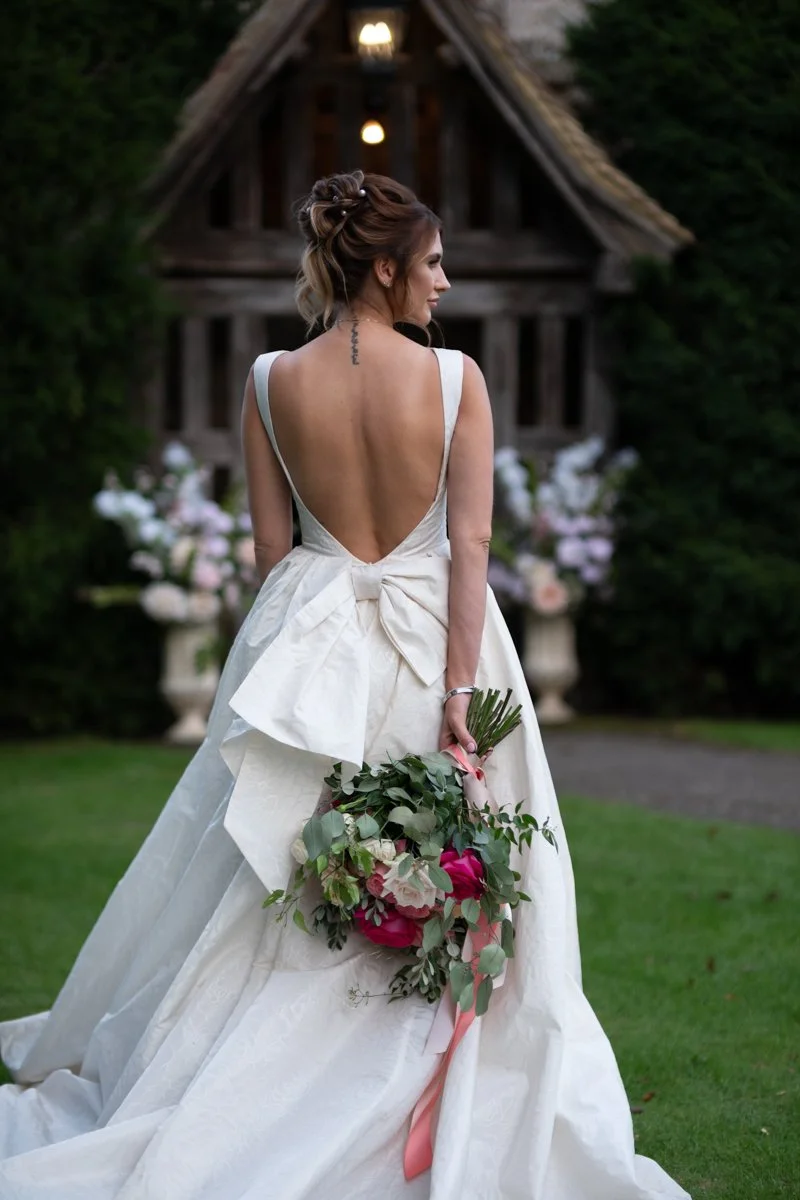 Bridal woman in a white wedding dress holding a bouquet of pink and white flowers outdoors with a rustic wooden chapel in the background.