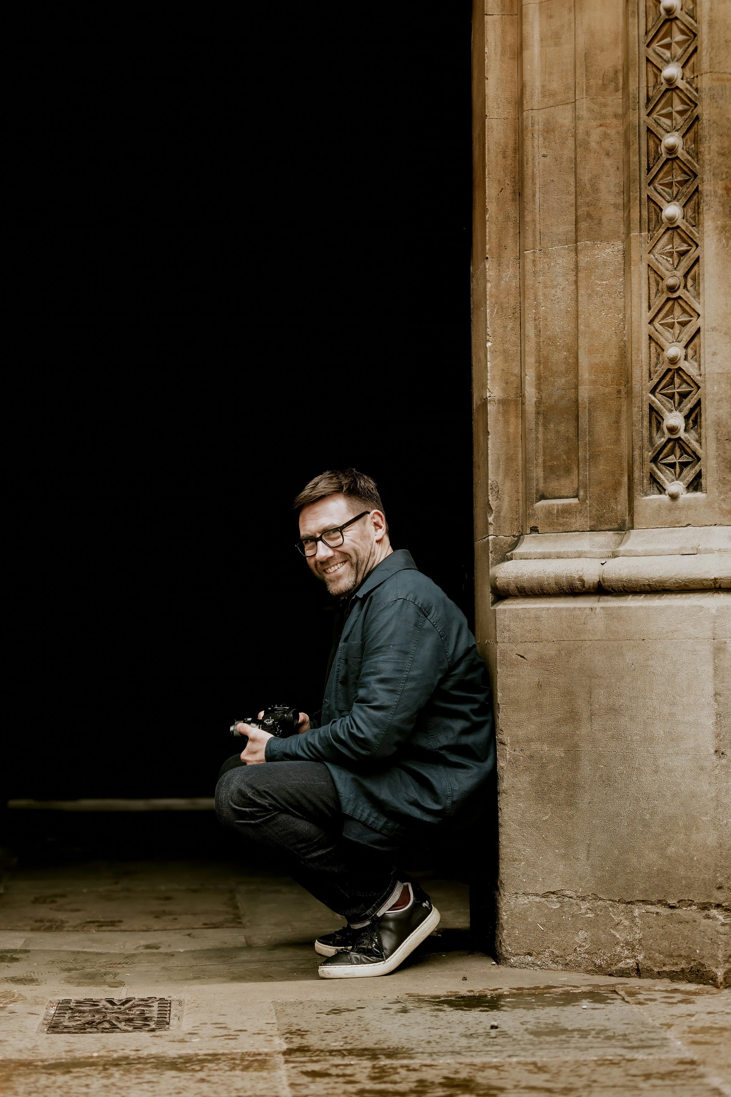 A smiling man wearing glasses, a dark jacket, and sneakers, crouching at the corner of a building with ornate architecture, holding a camera.