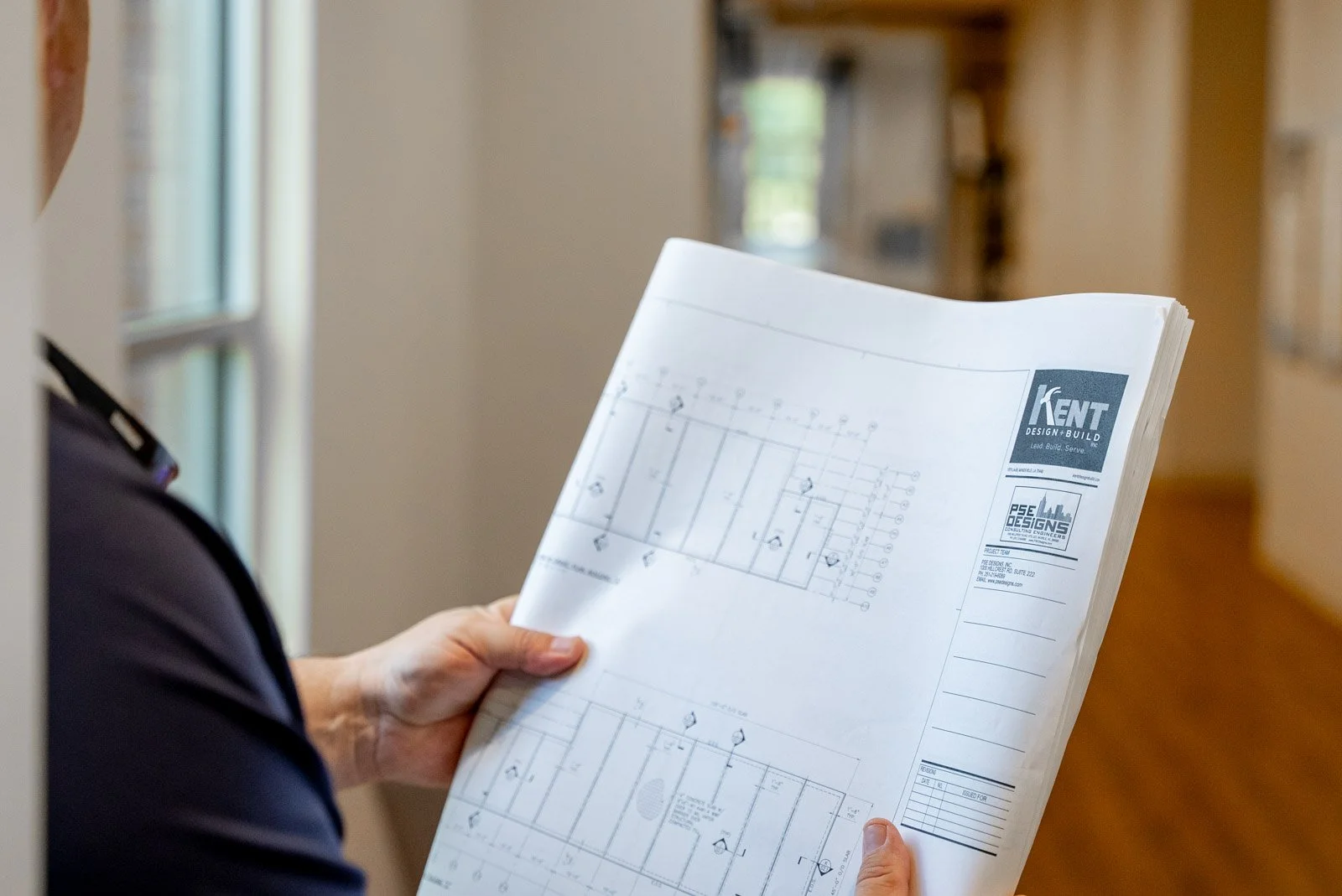 Person in a dark shirt holding architectural blueprint with company logos in a well-lit interior.