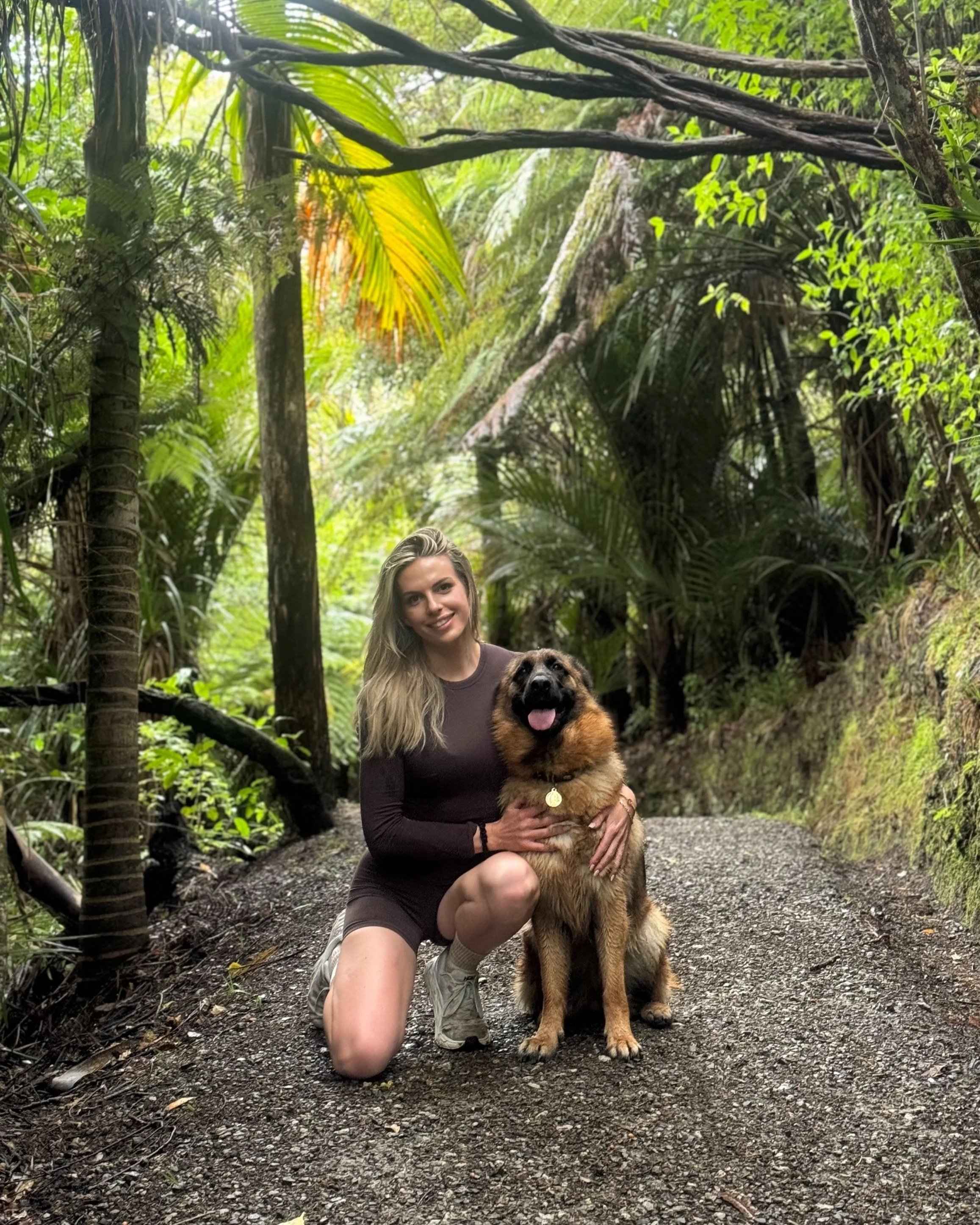 A woman with long blonde hair, wearing a dark long-sleeved shirt and white sneakers, kneeling on a dirt trail in a lush green jungle, holding a large, fluffy German Shepherd dog with a gold medal collar. The woman is smiling at the camera, and the dog appears happy with its tongue out.