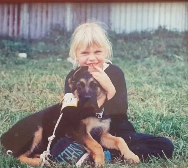 Young girl with blond hair smiling and hugging a large black and tan puppy sitting on grass in a backyard.