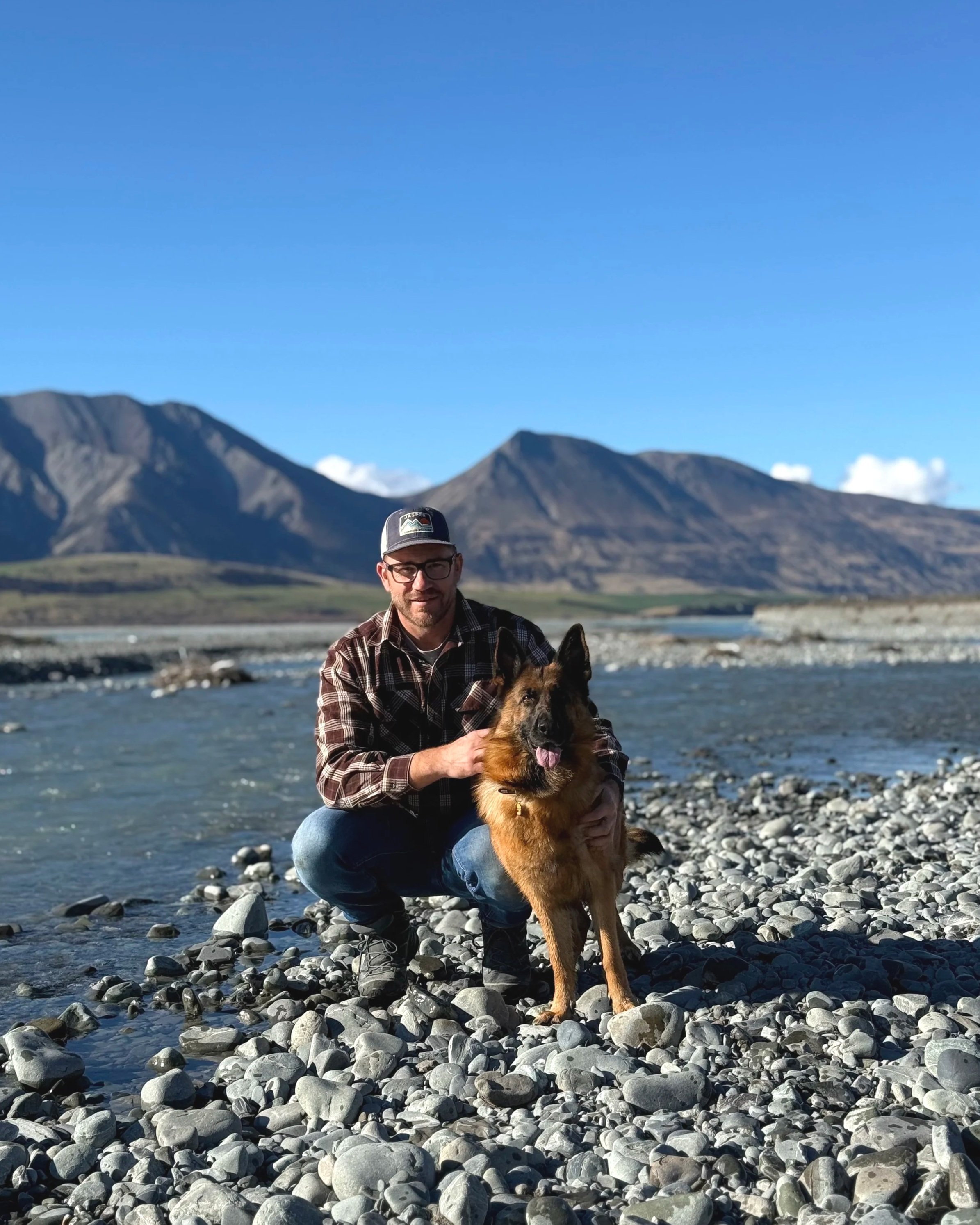 A man with a beard, glasses, and a cap crouching on a rocky riverbank, holding a German Shepherd dog. In the background are mountains under a clear blue sky.