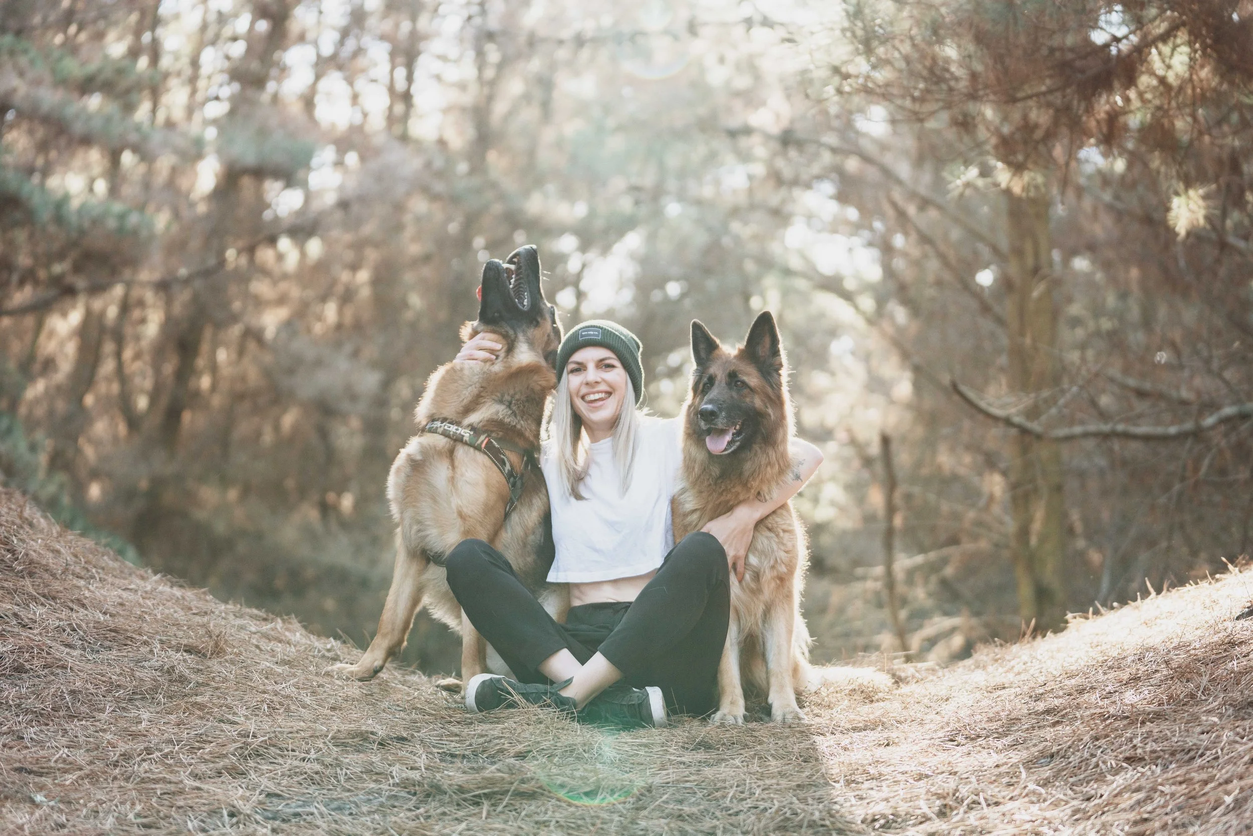 A woman sitting outdoors in a wooded area with two German Shepherd dogs, one of which is jumping and licking her face, while the other is sitting calmly.