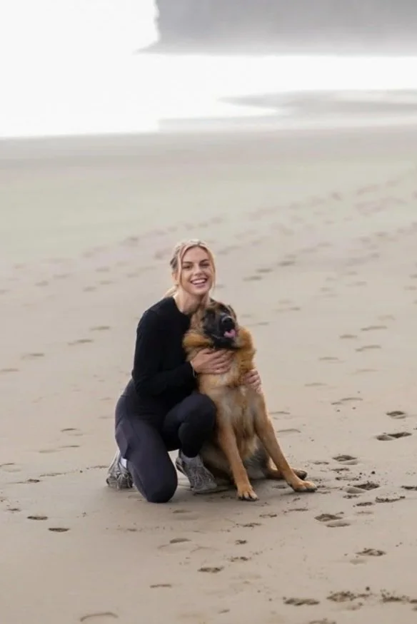 A woman kneeling on a beach, holding a large German Shepherd dog, both smiling. The beach has footprints on the sand, and the ocean is visible in the background under an overcast sky.