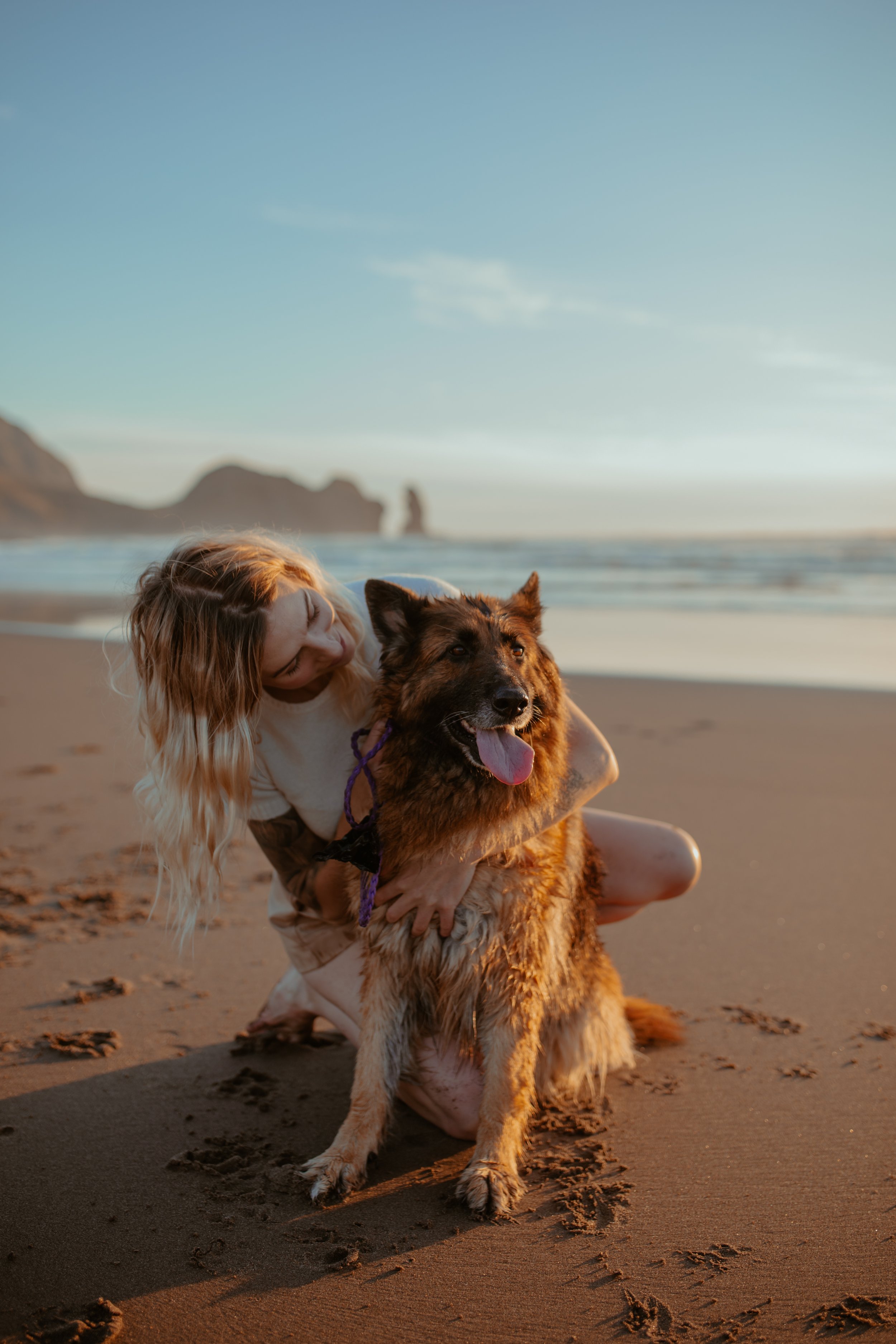 A woman crouching with a German Shepherd dog on a sandy beach during sunset, with cliffs and ocean in the background.
