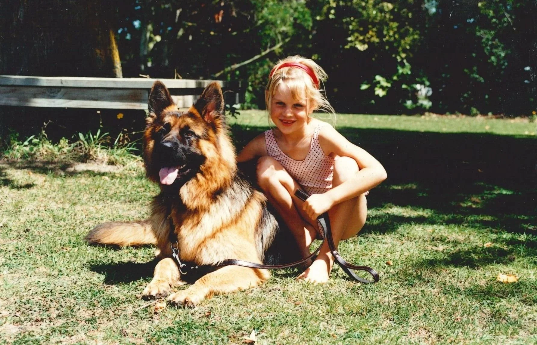 A young girl with blonde hair and a pink headband, wearing a polka dot dress, squatting on the grass and smiling next to a large German Shepherd dog lying on the ground. The girl is holding the dog's leash. There is green grass, trees, and a wooden structure in the background.