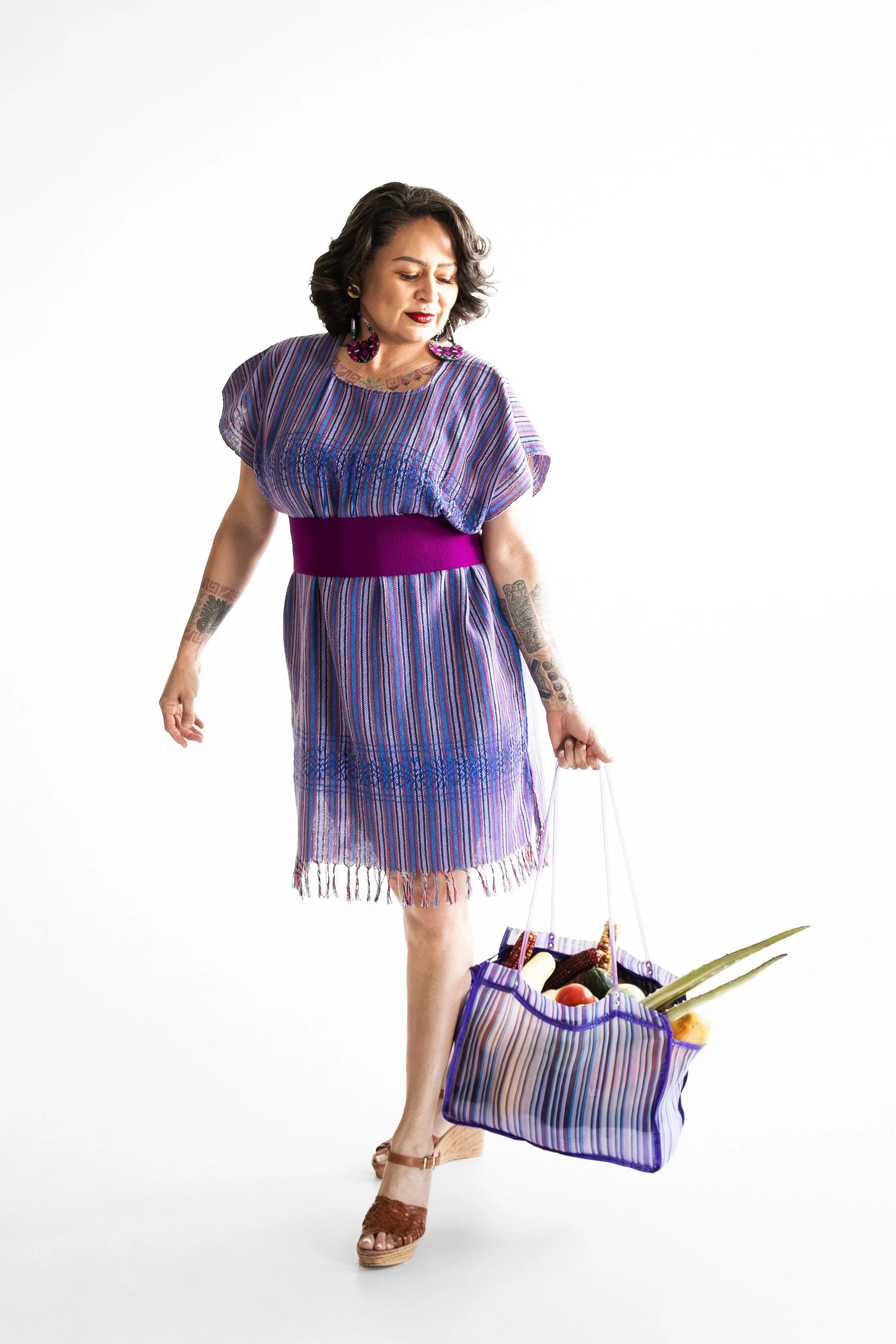 Woman in colorful striped dress holding a striped shopping bag filled with fruits and vegetables, standing on a plain white background.