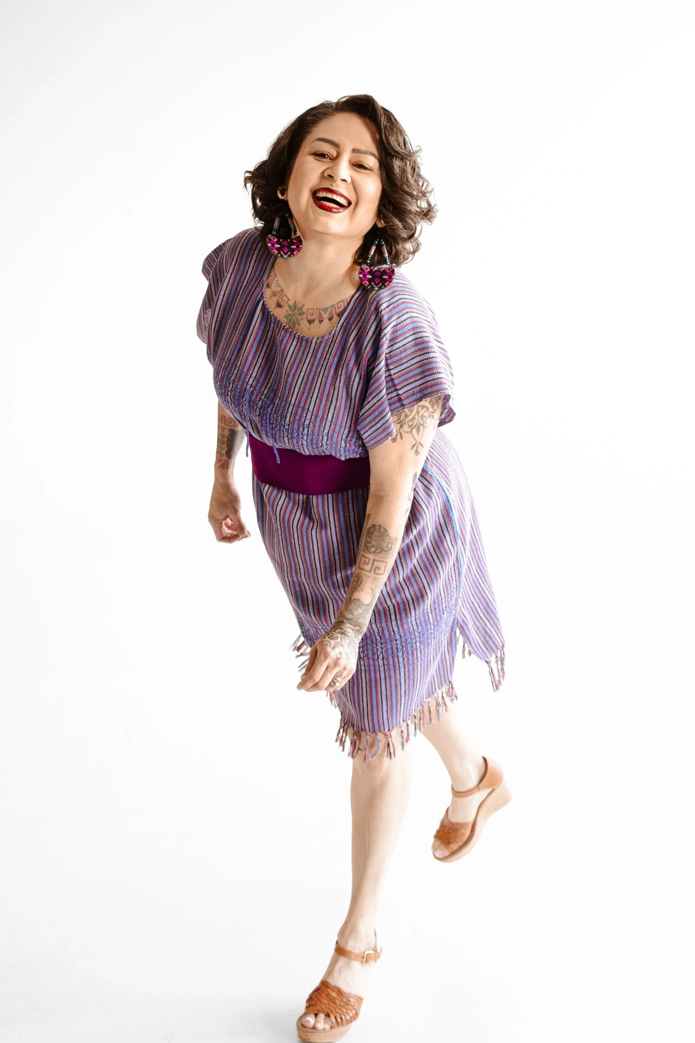 A woman with dark curly hair, tattoos, and red lipstick wearing a colorful striped dress with fringes, large beaded earrings, and sandals, smiling and striking a playful pose against a plain white background.